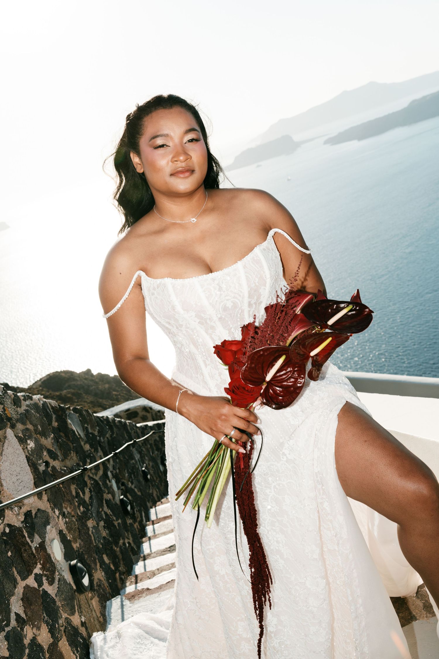 Bride in white lace dress holding red floral bouquet on scenic Santorini terrace overlooking blue sea.