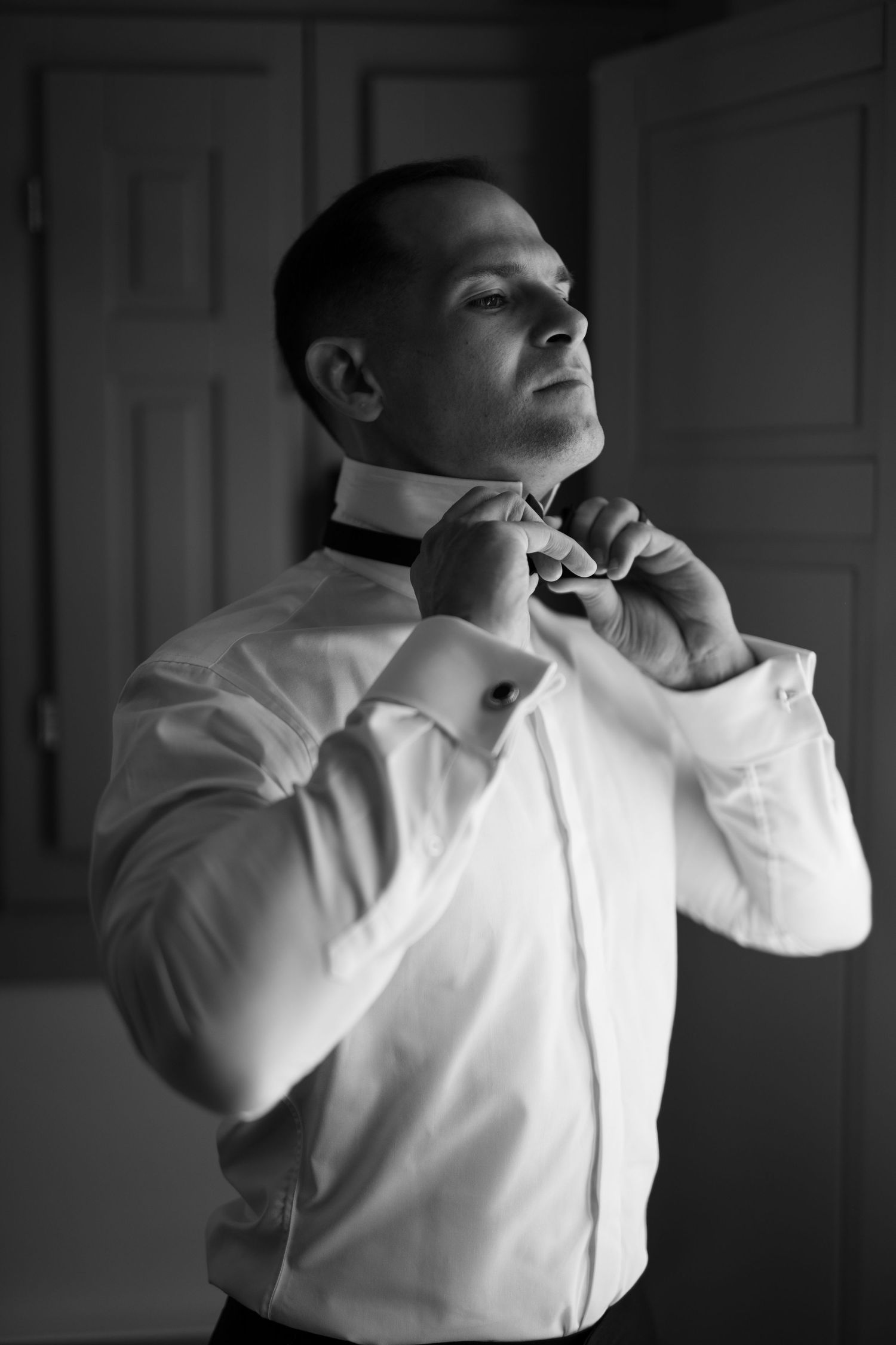 Black and white photo of a man in a white dress shirt adjusting his bow tie, looking upward confidently.