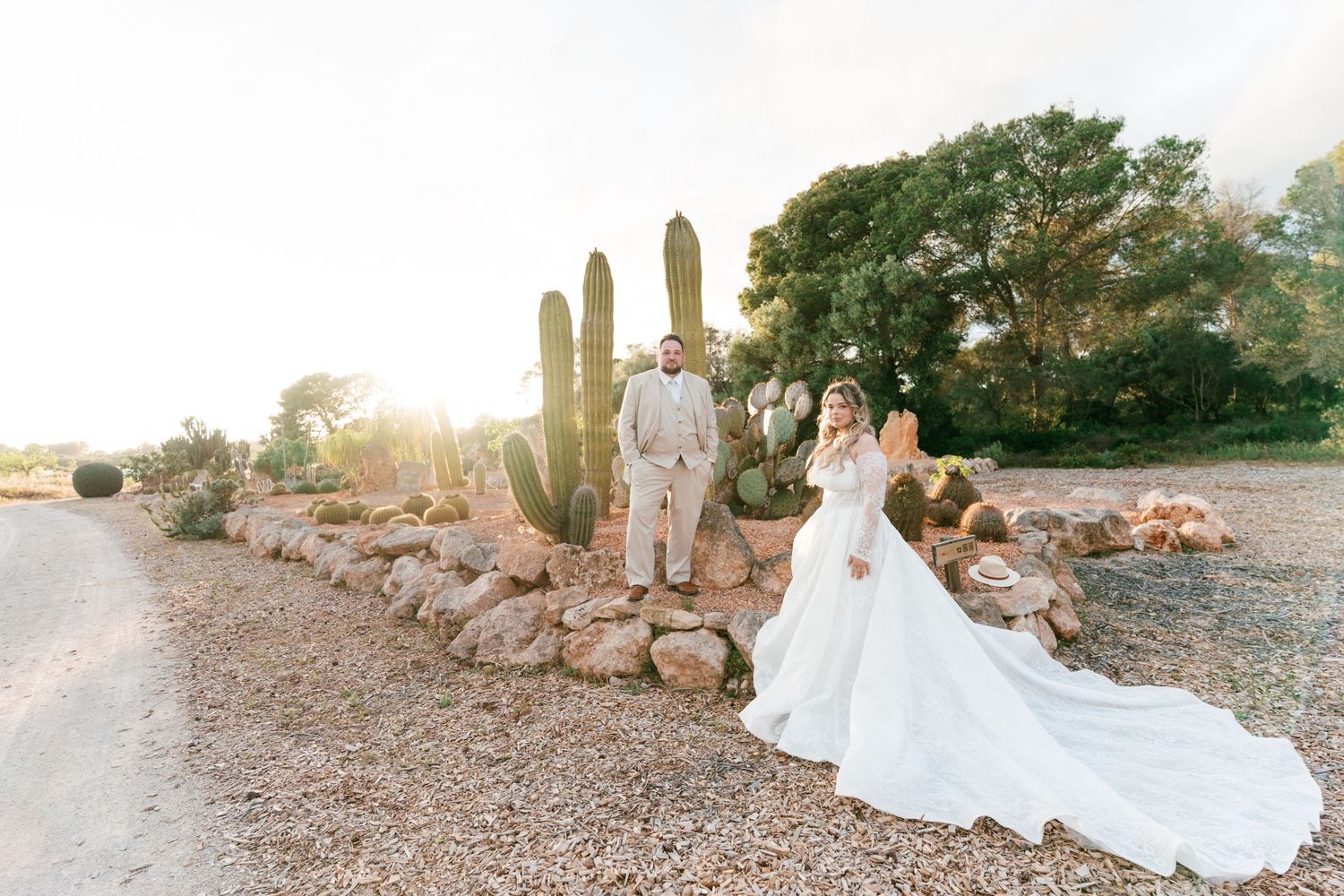 Bride and groom posing near tall cacti and rocks in a desert landscape with warm golden sunlight in Mallorca.