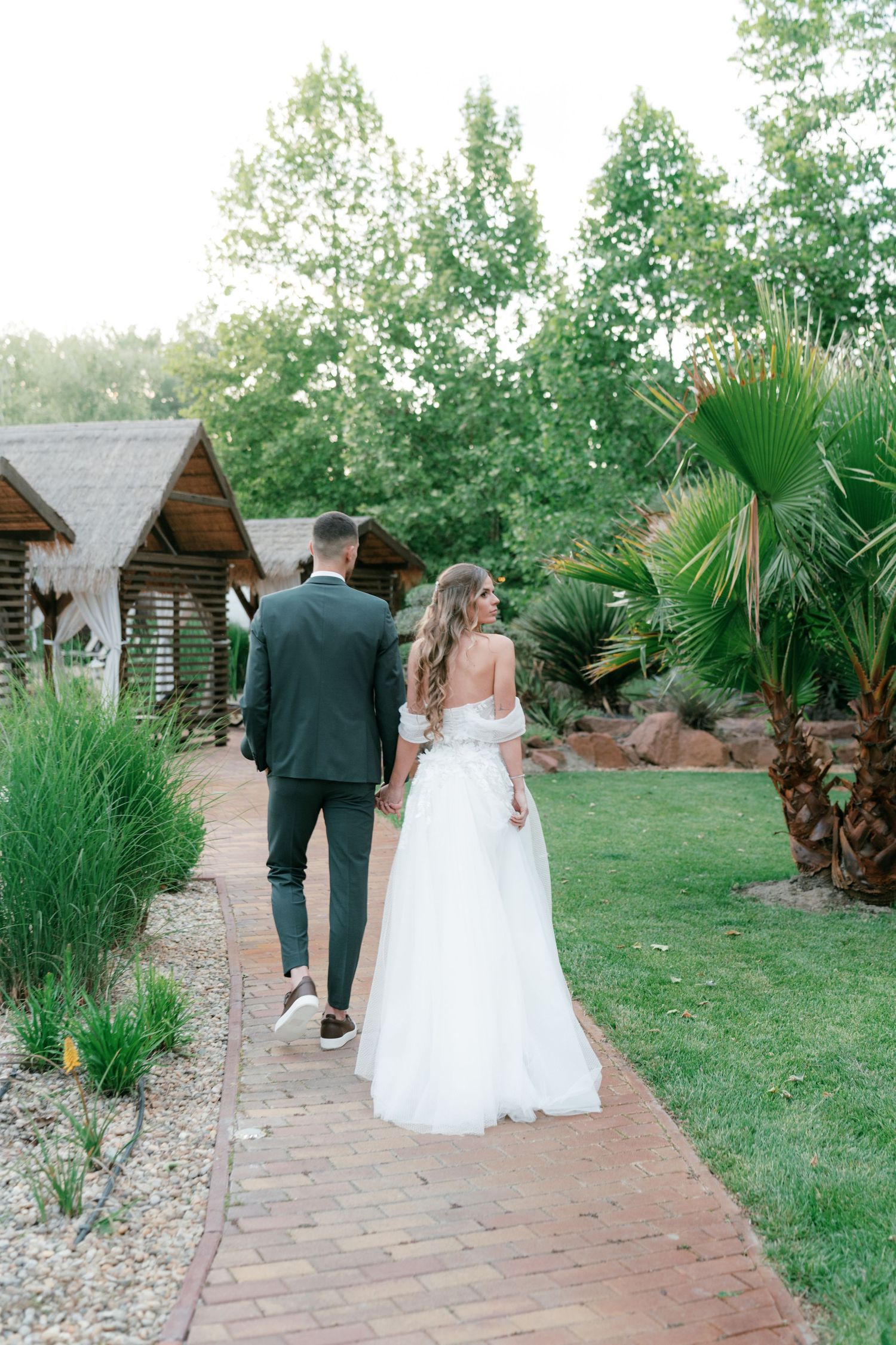 Bride and groom walk hand in hand along brick path through lush tropical wedding venue. Mallorca wedding photographer and filmmaker.