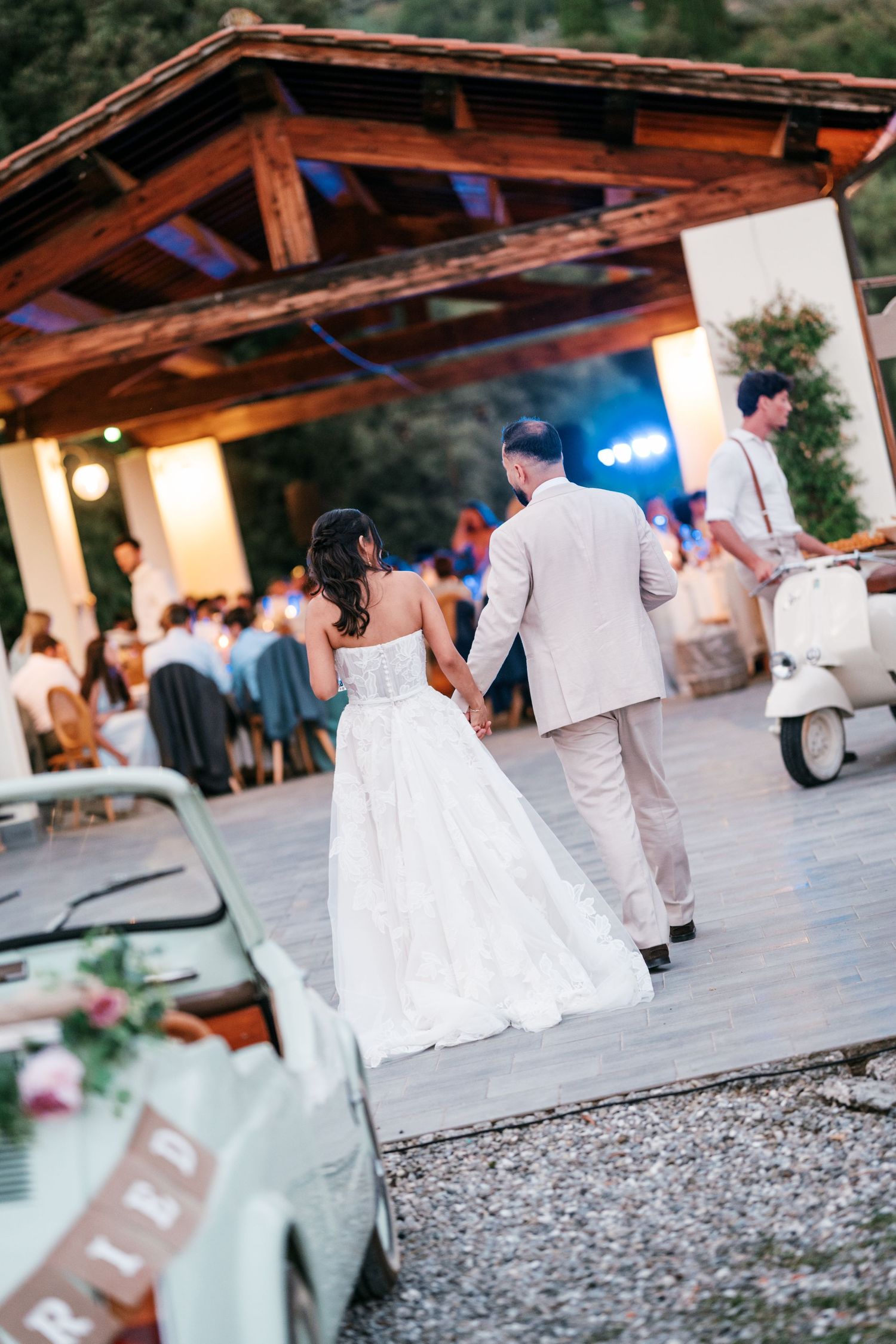 Bride in a white gown and groom in a beige suit walk hand in hand toward an outdoor wedding reception with a vintage car and scooter.