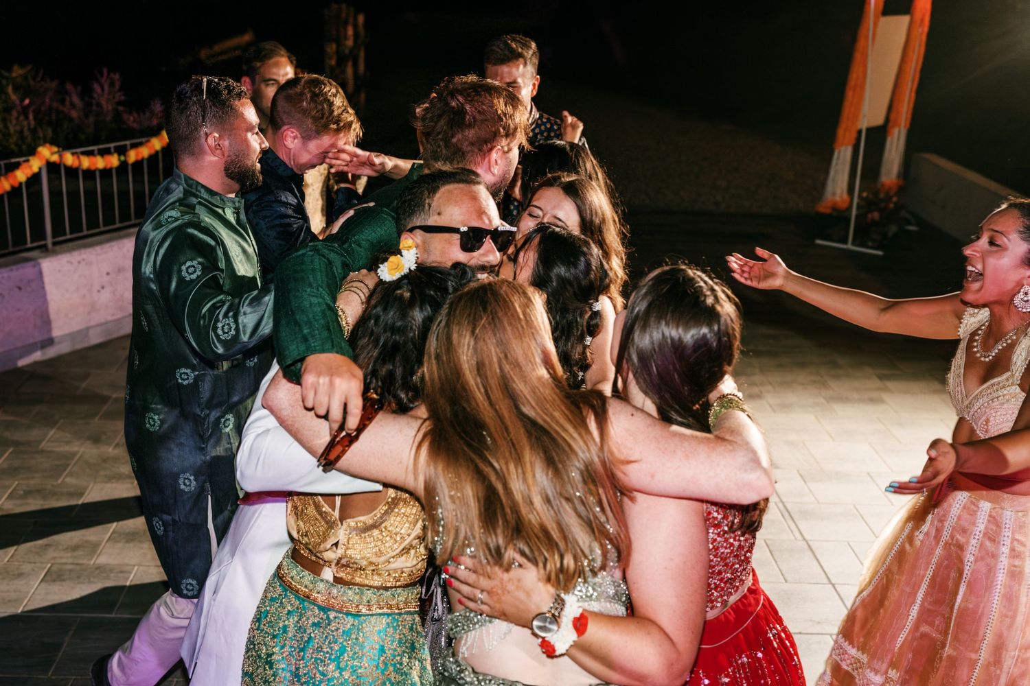 Wedding guests in colorful traditional Indian attire gather in a joyful group hug on an outdoor dance floor at night.
