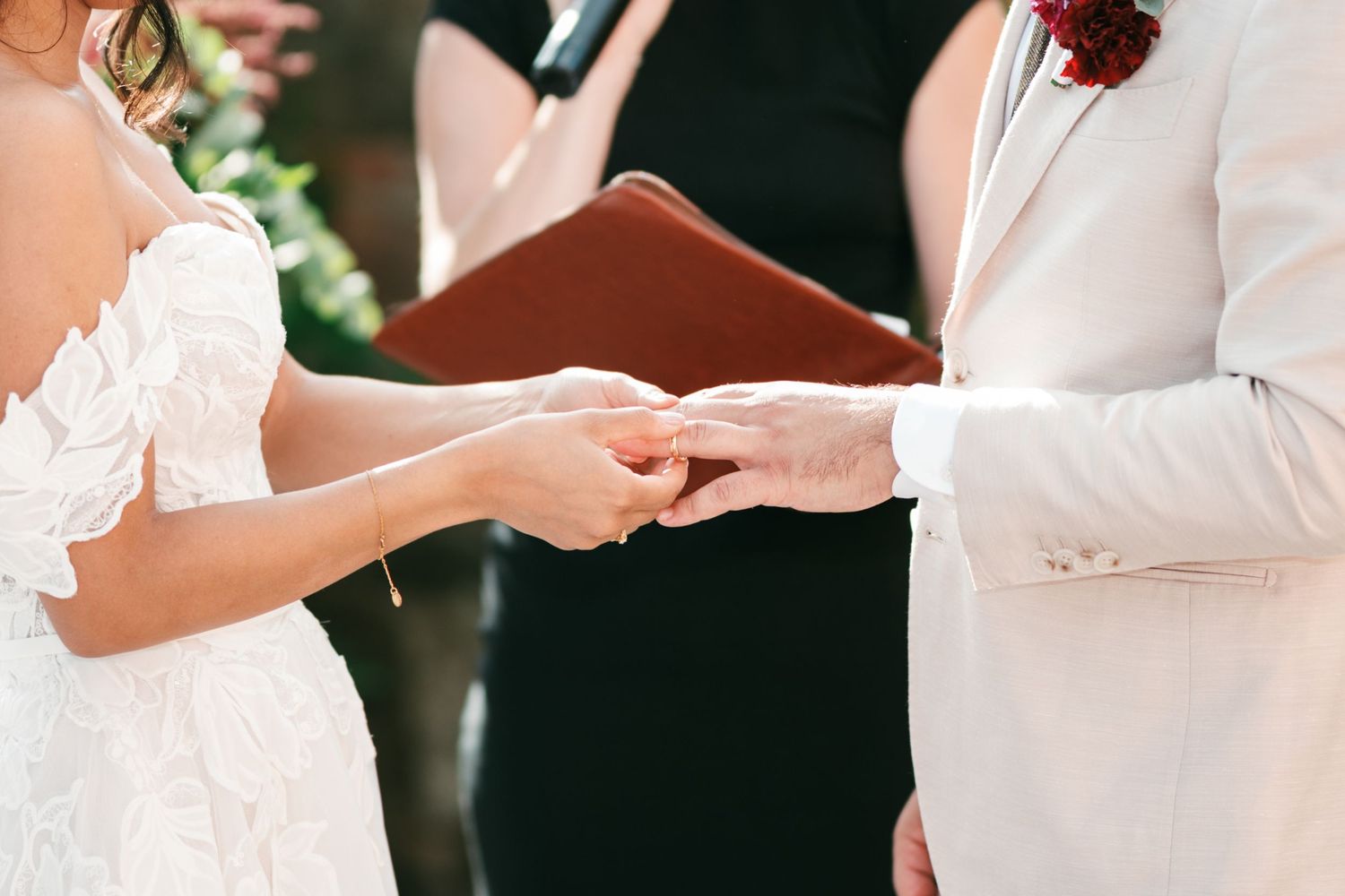 Bride in a white lace gown and groom in a light suit hold hands during a wedding ceremony while an officiant holds a leather book.