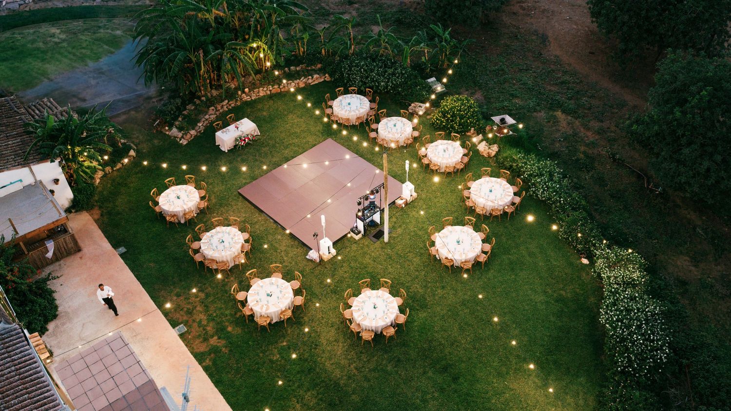 Aerial view of a garden wedding reception with round tables, a wooden dance floor and string lights on a green lawn at dusk.