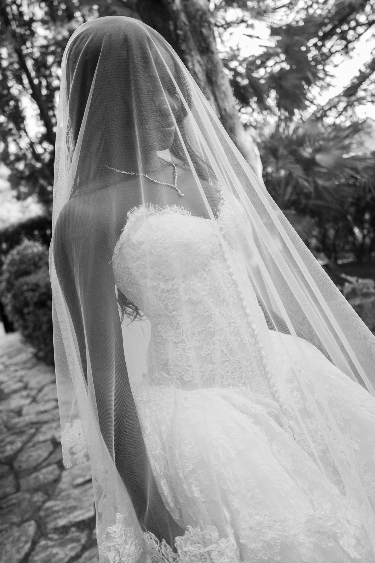 Black and white photo of a bride in a lace strapless wedding gown with a sheer veil draped over her head, standing on a stone garden path.