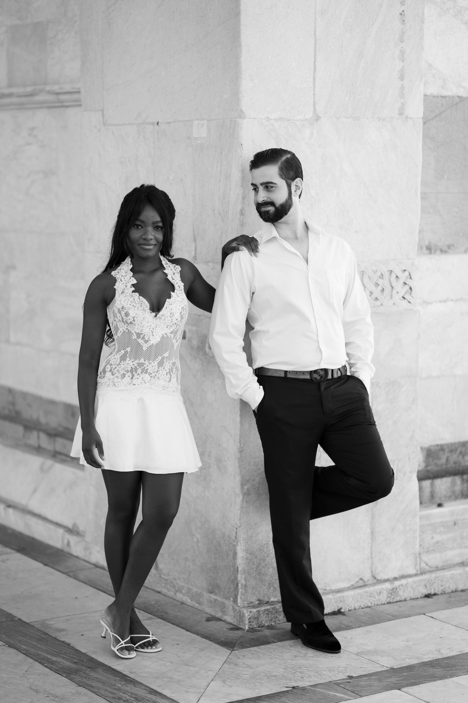 Black and white portrait of a smiling couple posing against a marble wall, she in a lace dress and he in a shirt and dress pants.