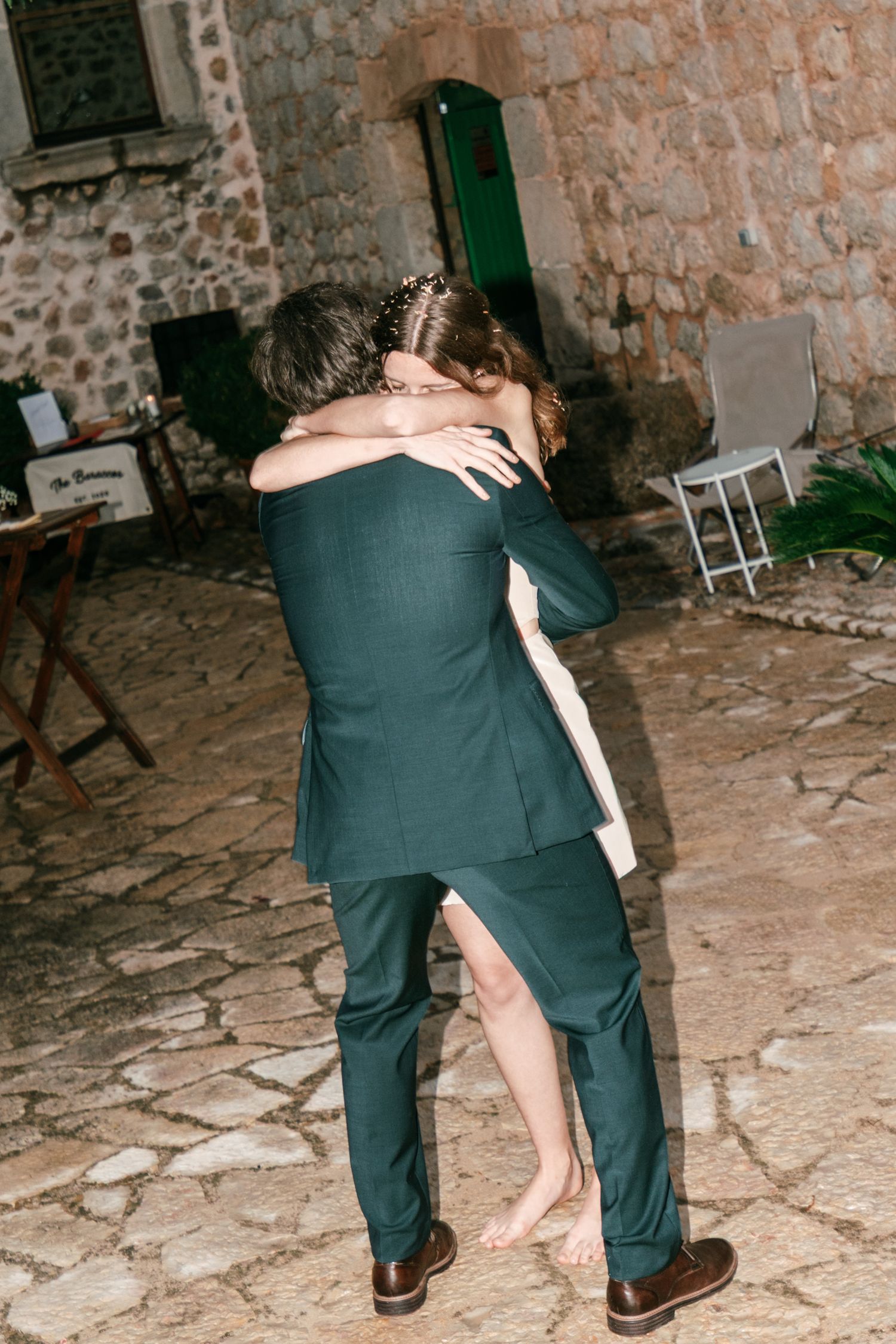 A groom in a green suit embraces a barefoot bride in a short white dress on a stone courtyard at a rustic wedding.