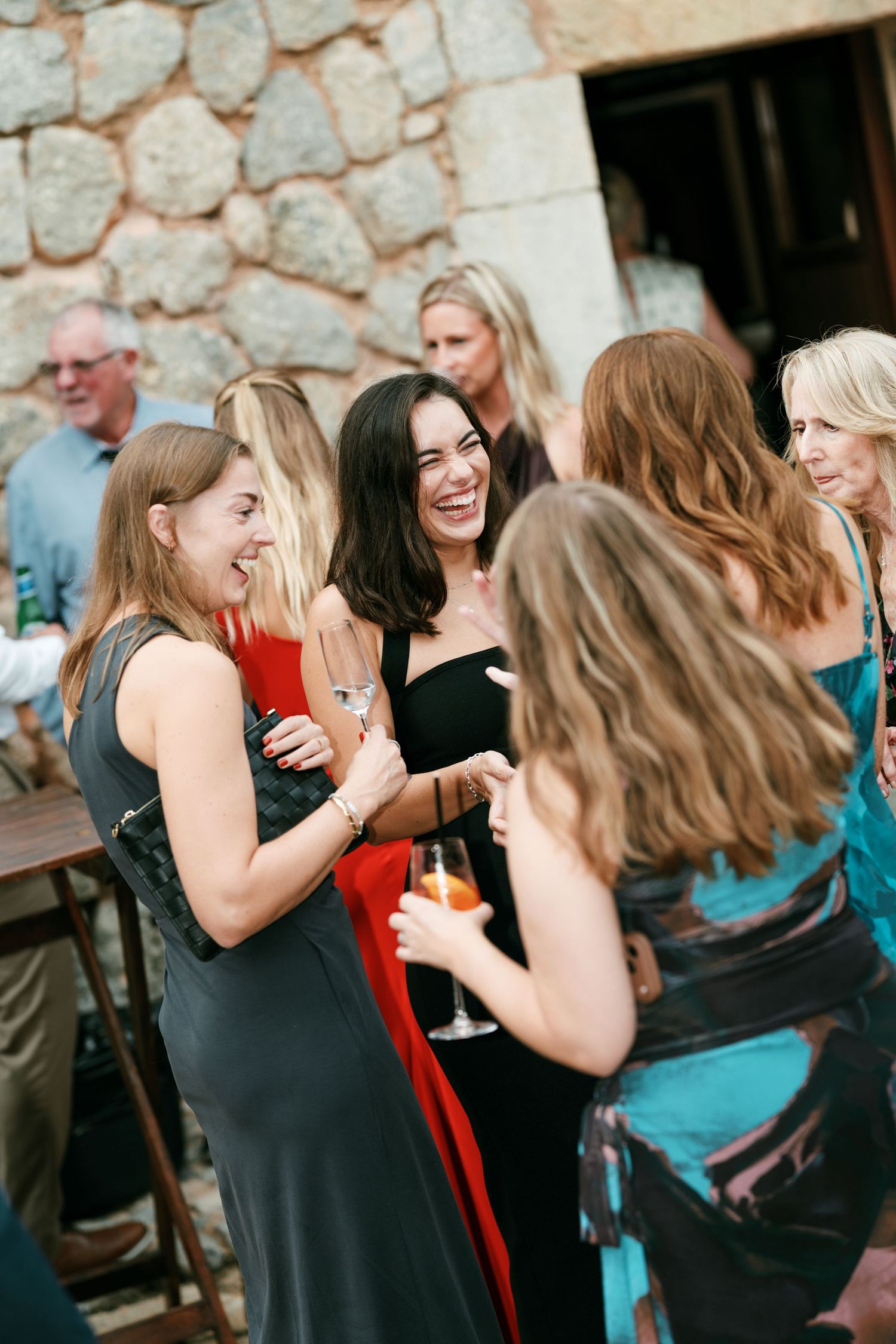 Smiling women in elegant dresses laugh and hold drinks while mingling at a lively outdoor wedding reception beside a stone wall.