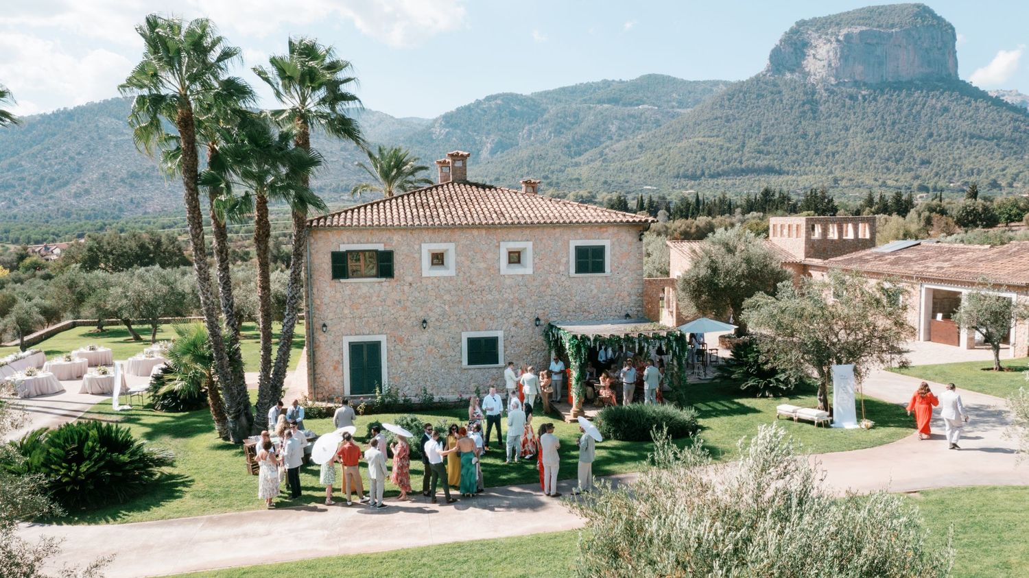 Guests gather outside a rustic stone villa surrounded by palm trees and mountains for an outdoor event in Mallorca.