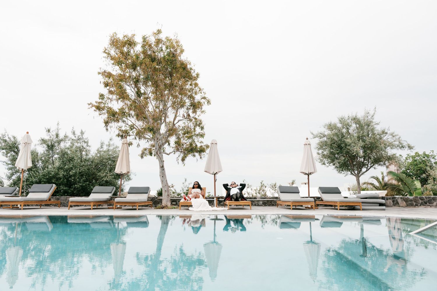 Elegant resort pool with lounge chairs, white umbrellas, and lush trees reflecting in calm water on an overcast day.