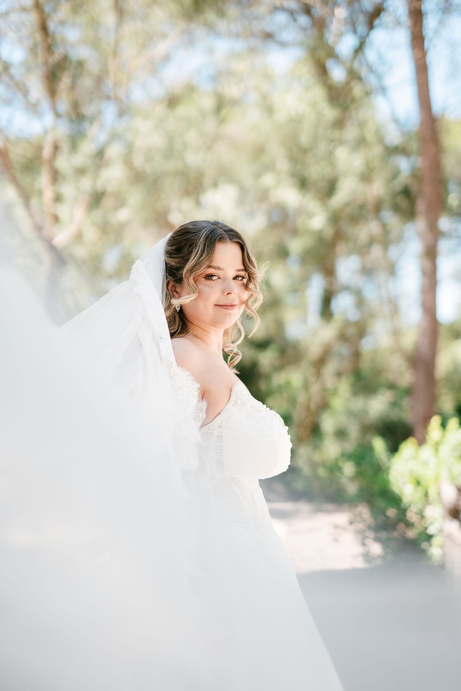 Bride in white gown and flowing veil smiling outdoors with lush green trees in soft sunlit background.
