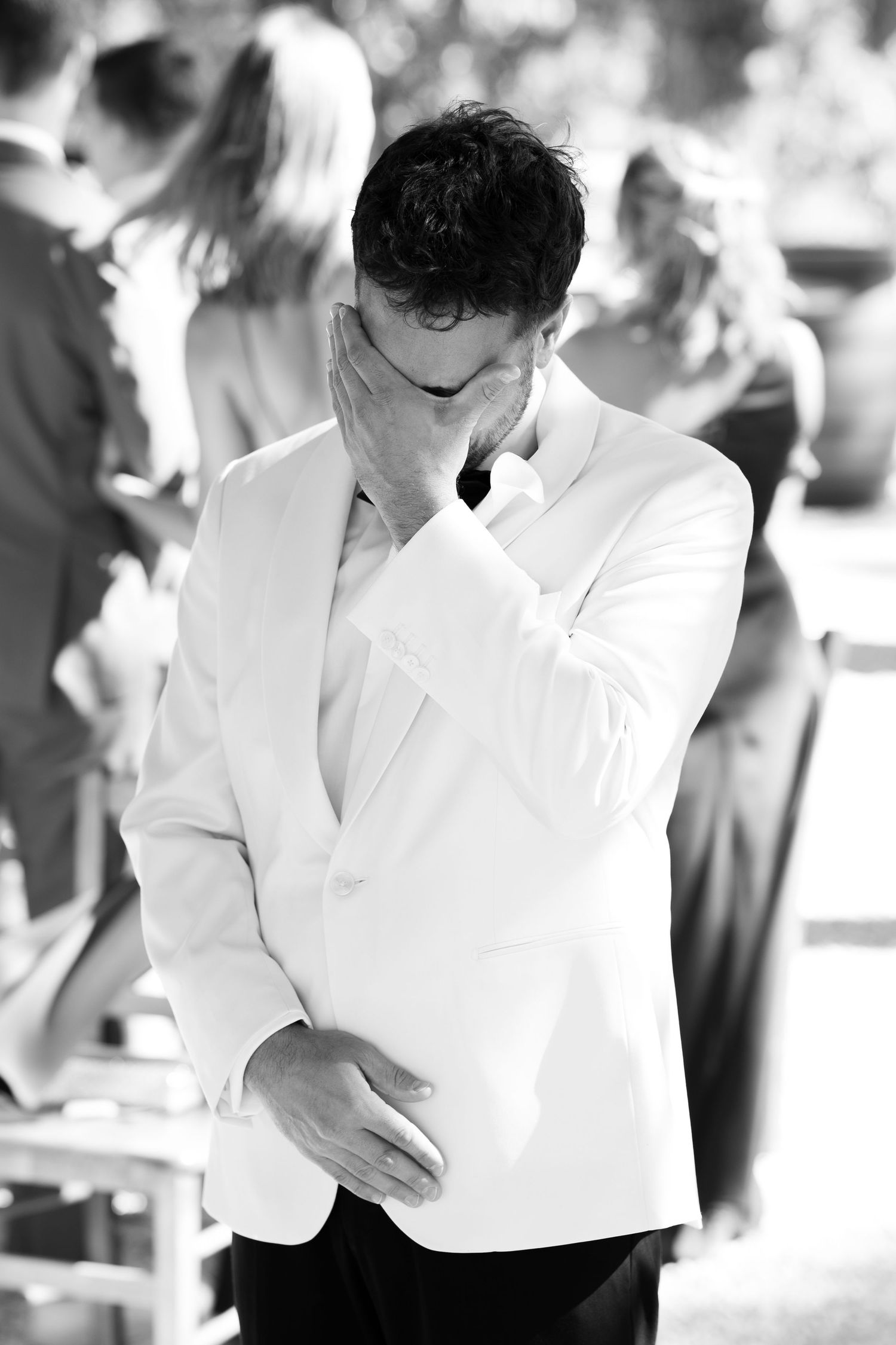 Black and white photo of emotional groom in white tuxedo jacket wiping tears during outdoor wedding ceremony.