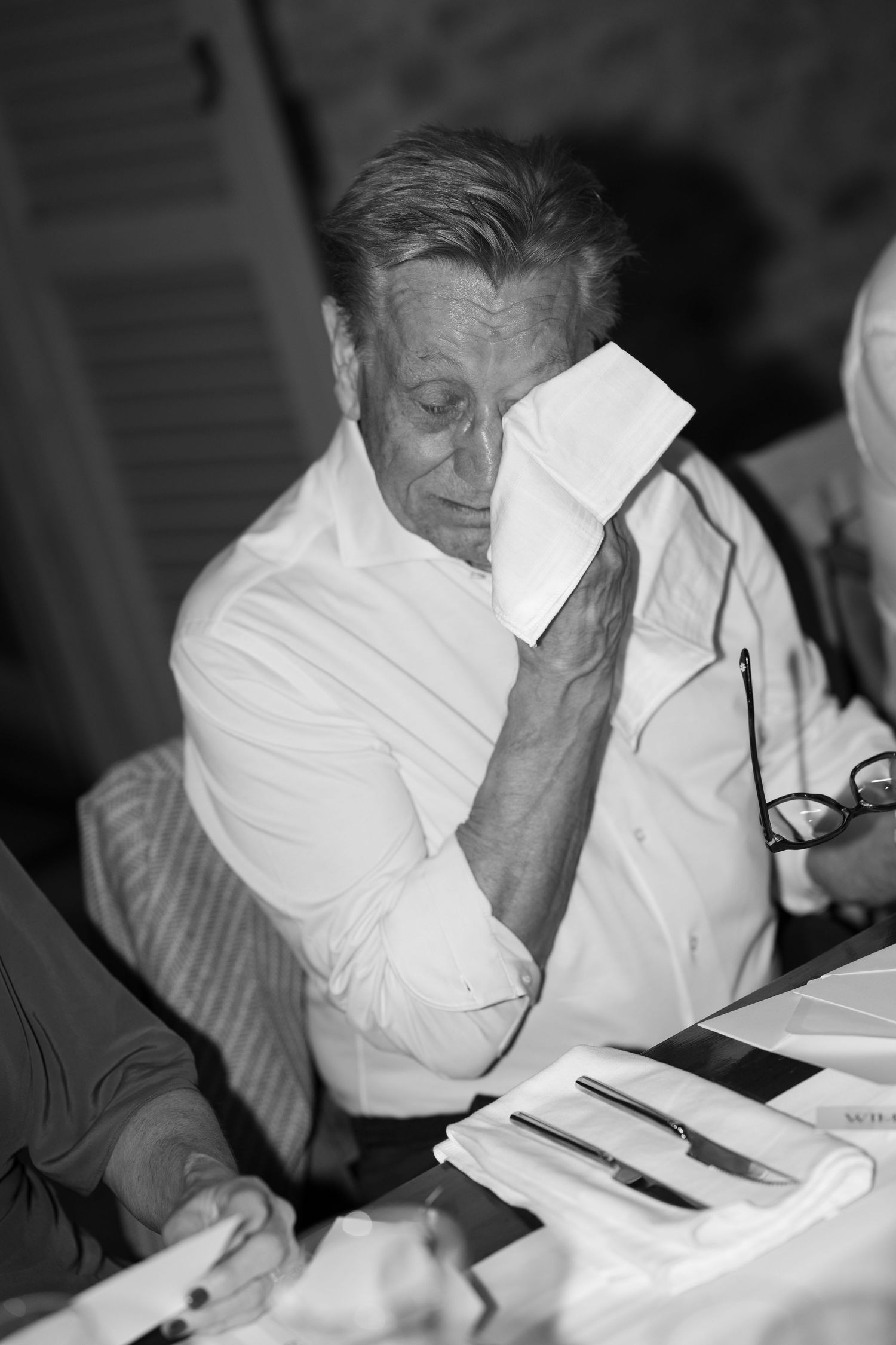 Black and white photo of an elderly man in a white shirt wiping his face with a napkin at a dining table.