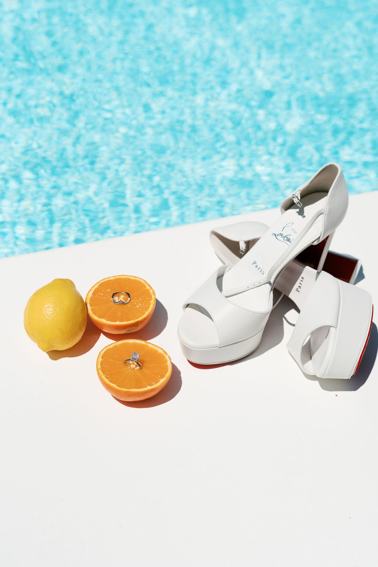 White platform heels with red soles beside sliced oranges and lemon with rings on white surface near blue pool.
