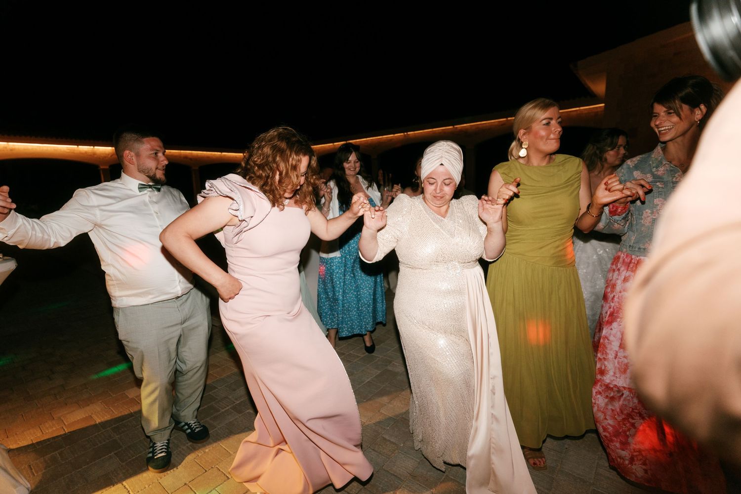 Guests dancing joyfully at an outdoor wedding reception at night, wearing elegant formal gowns in pink, white, and green in Mallorca.