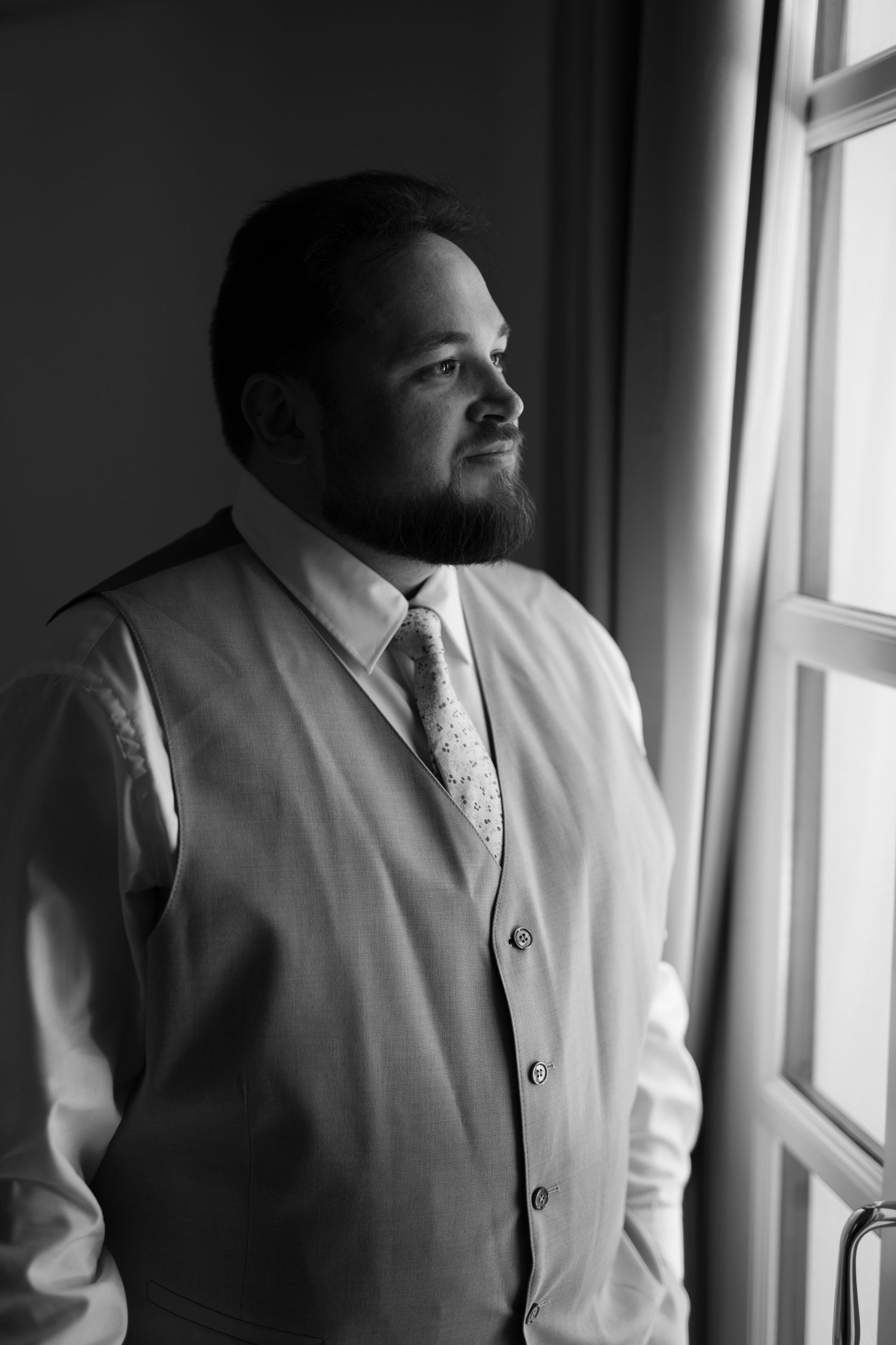 Black and white portrait of a bearded man in a vest and tie gazing thoughtfully out a window in soft natural light in Mallorca.