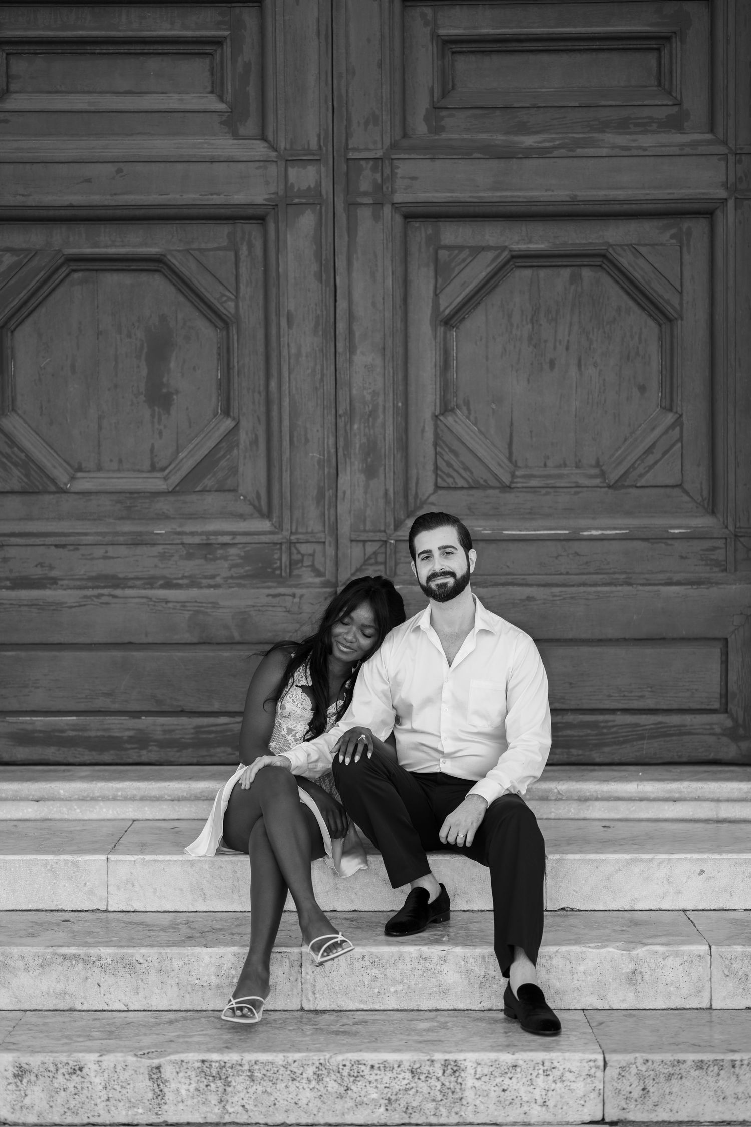 Black and white photo of a couple sitting on stone steps in front of an ornate wooden door, her head resting on his shoulder.