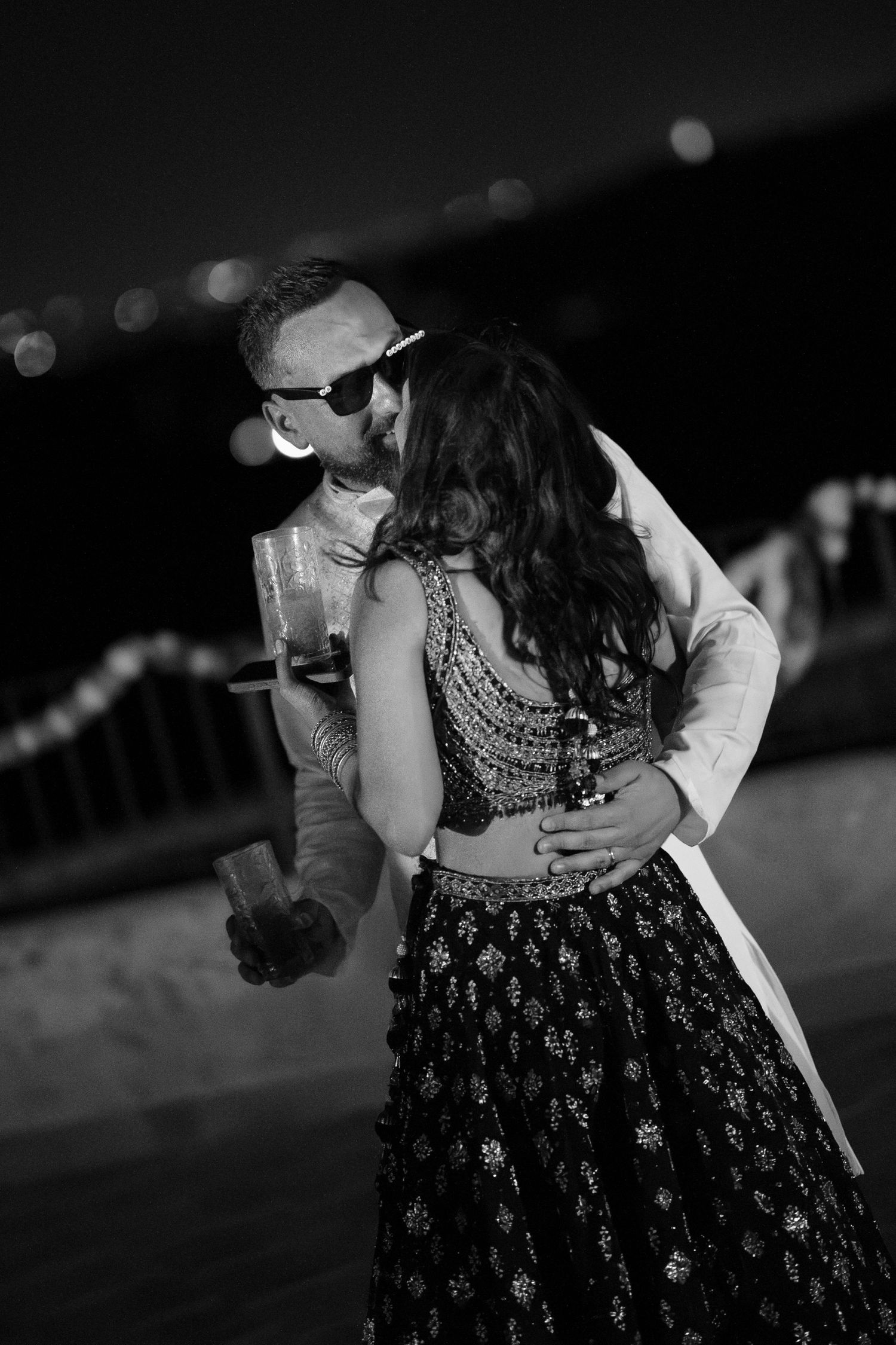 Black and white photo of a groom in sunglasses embracing a bride in an embroidered lehenga during an evening celebration.