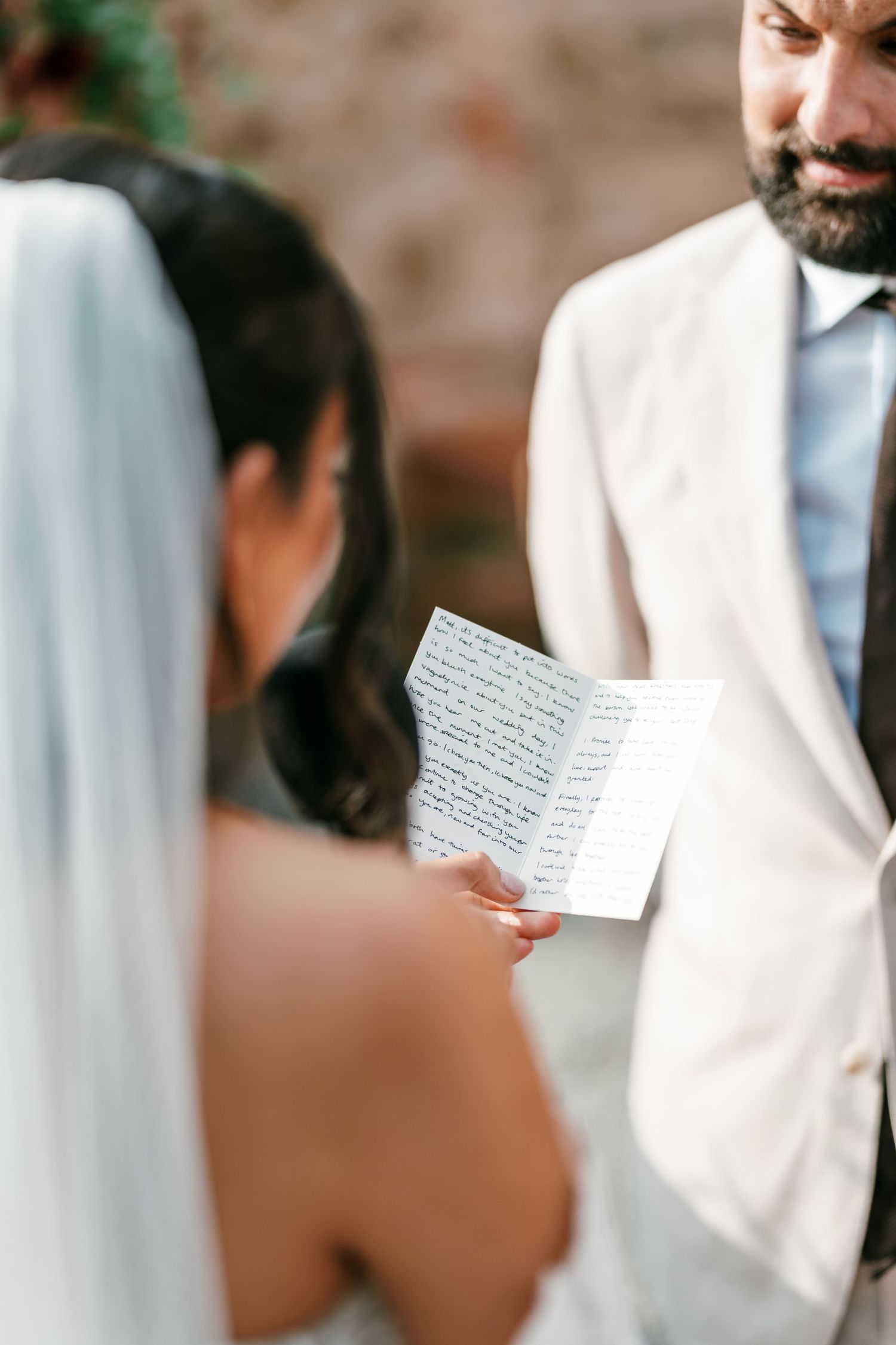 Bride in a white veil reads handwritten vows from a folded note to a bearded groom in a cream suit during an outdoor wedding ceremony.