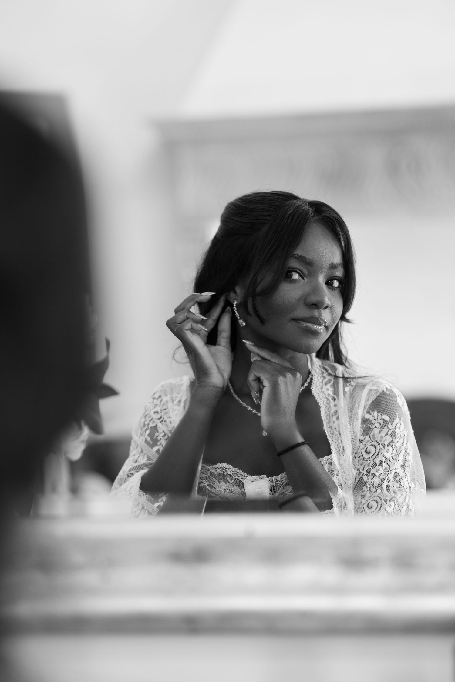 Black and white portrait of a woman in a lace robe adjusting her earring while smiling at her reflection in a mirror.