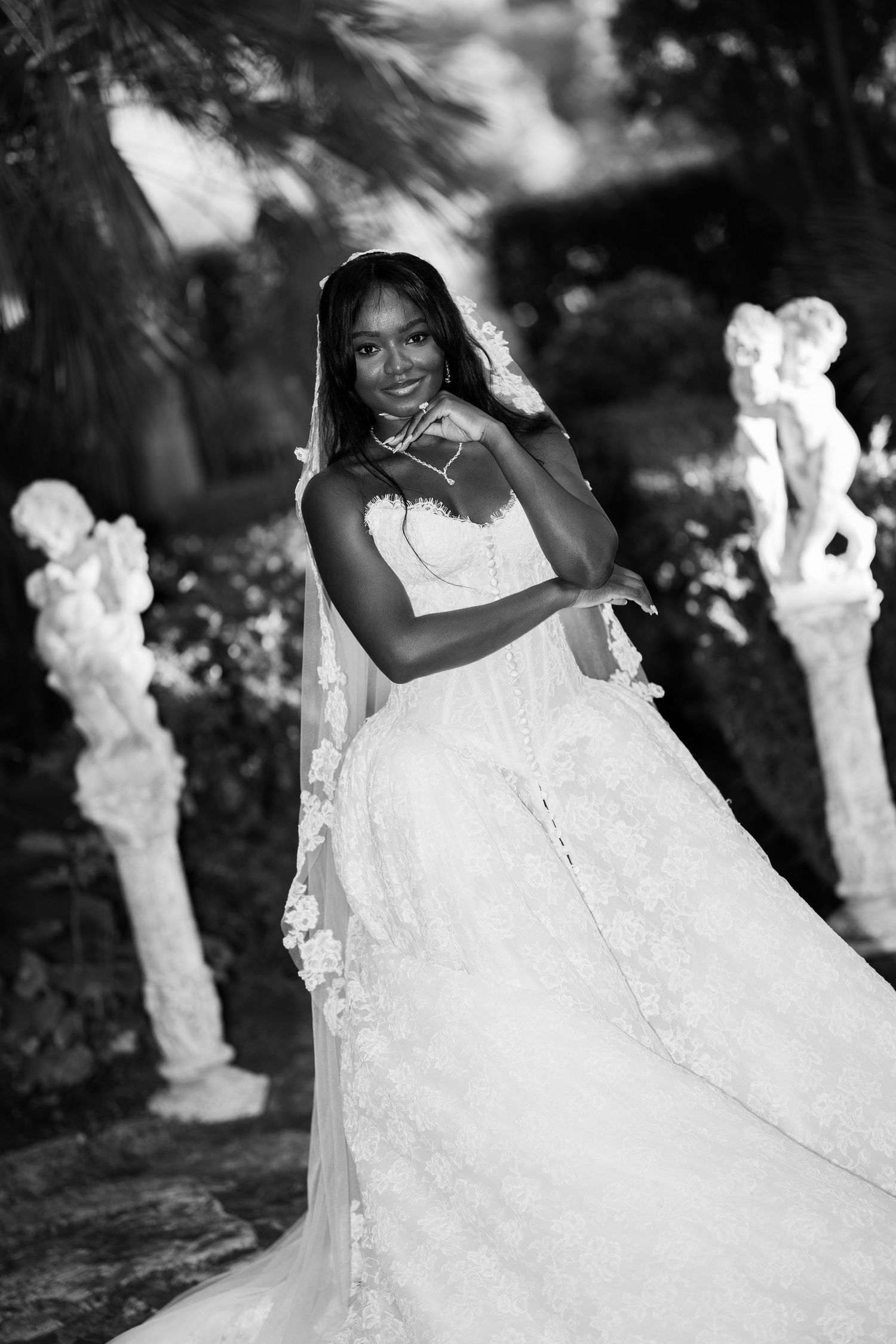 Black and white portrait of a smiling bride in a lace corset gown and floral veil posing among stone cherub statues in a garden.