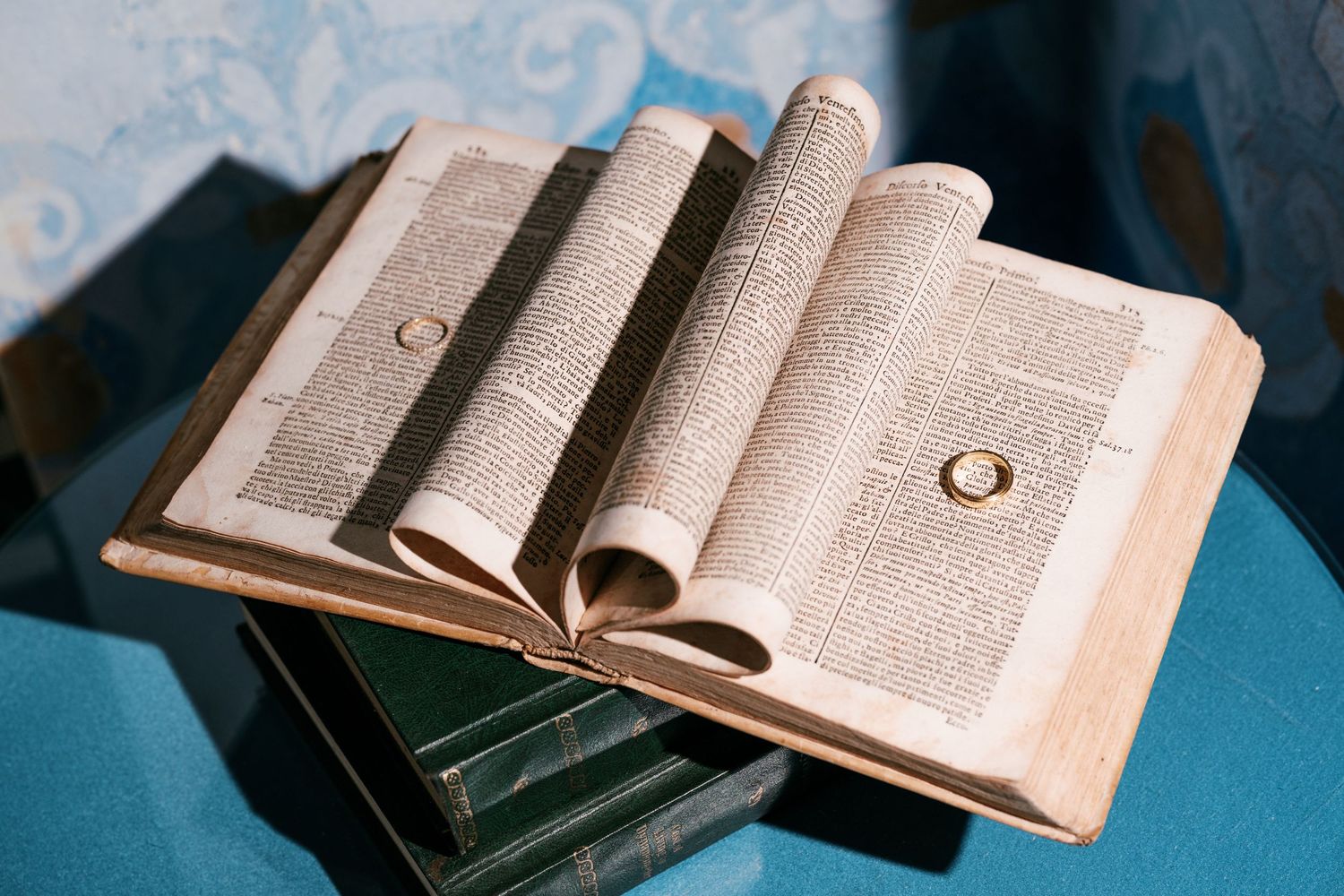 Open antique book with yellowed pages and two gold rings resting on its curled text, placed on a stack of old books.