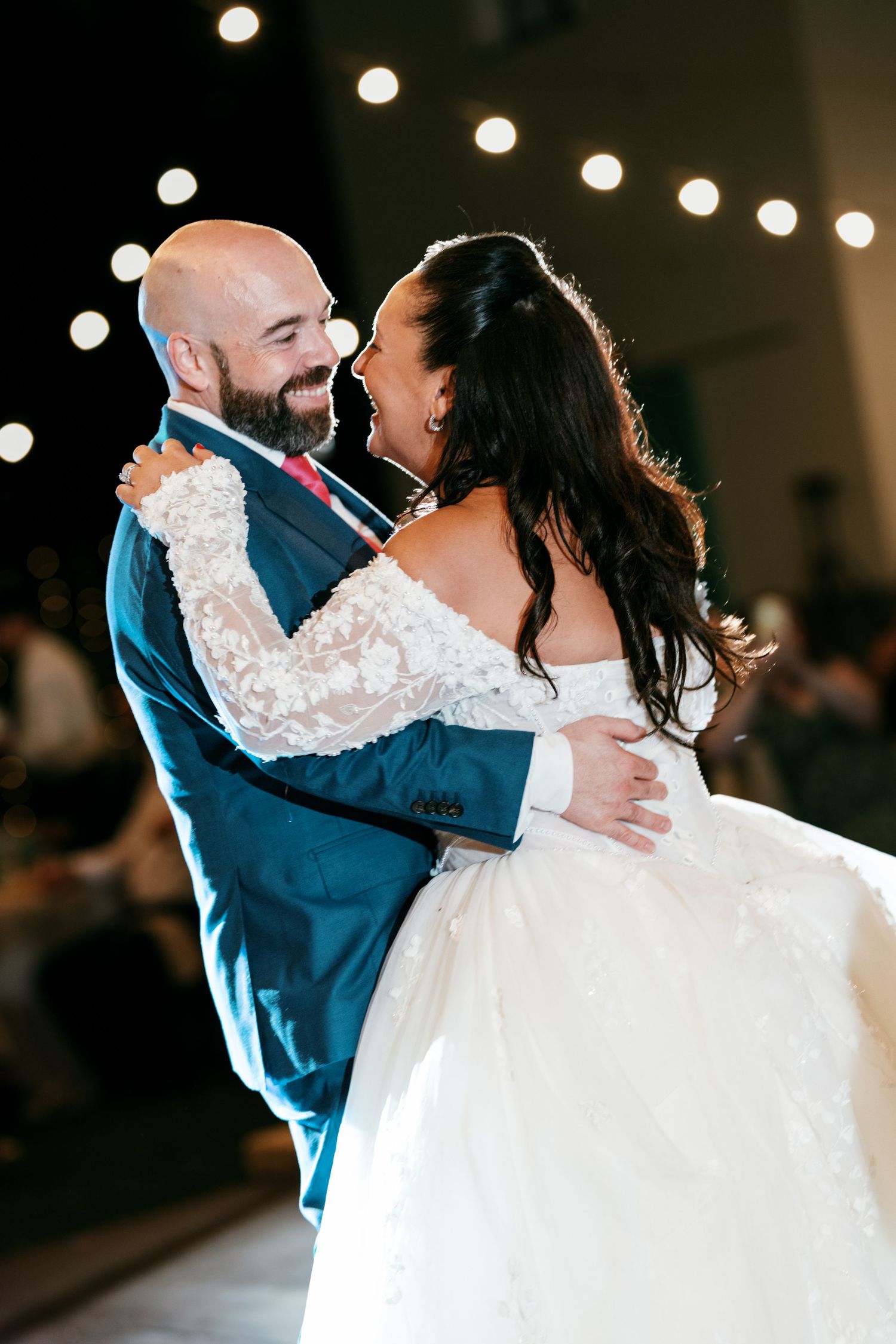 A smiling groom in a teal suit embraces his bride in an off-shoulder lace gown while dancing under warm string lights.