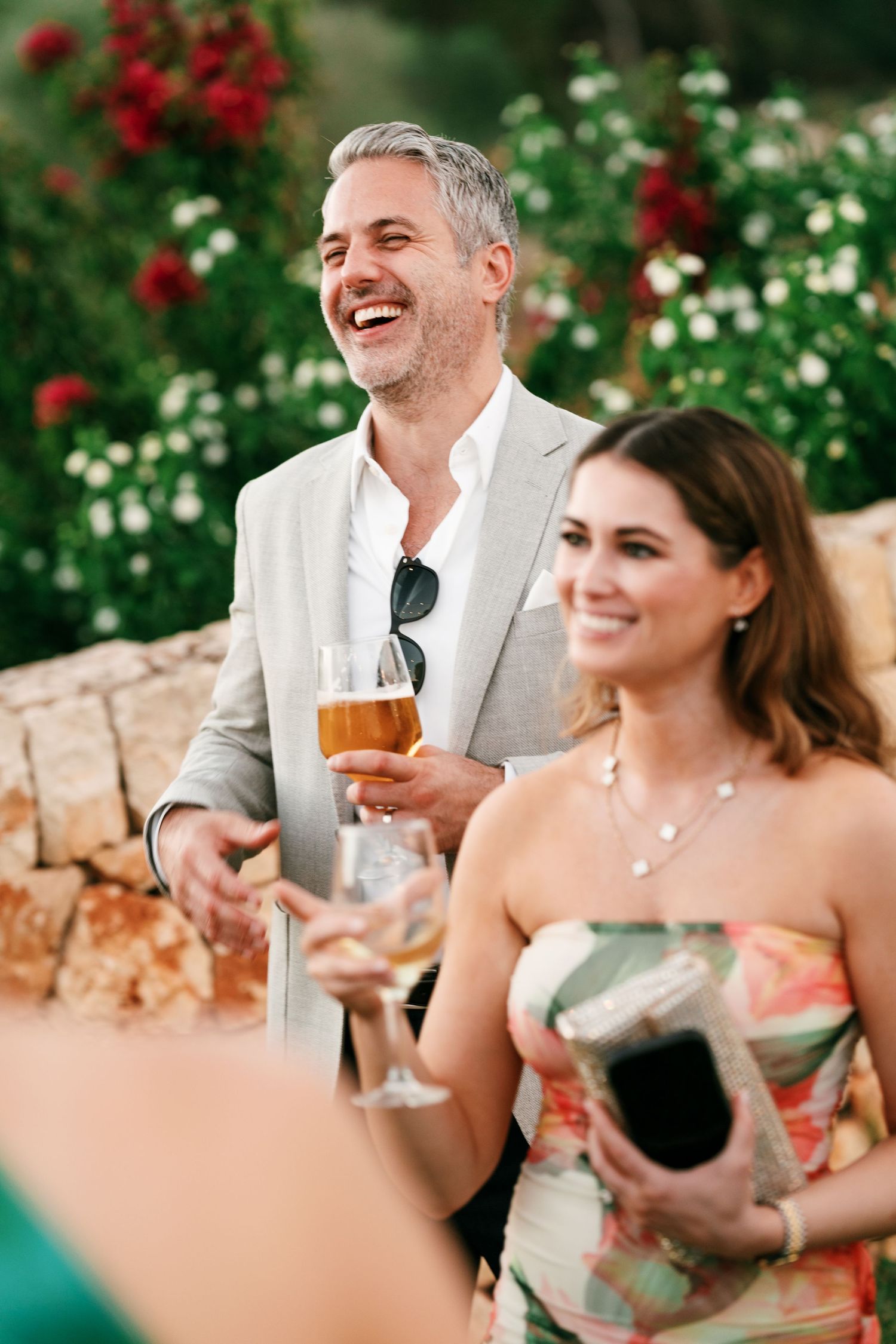 A laughing man in a light blazer holds a glass of beer beside a smiling woman in a floral dress at an outdoor garden celebration.