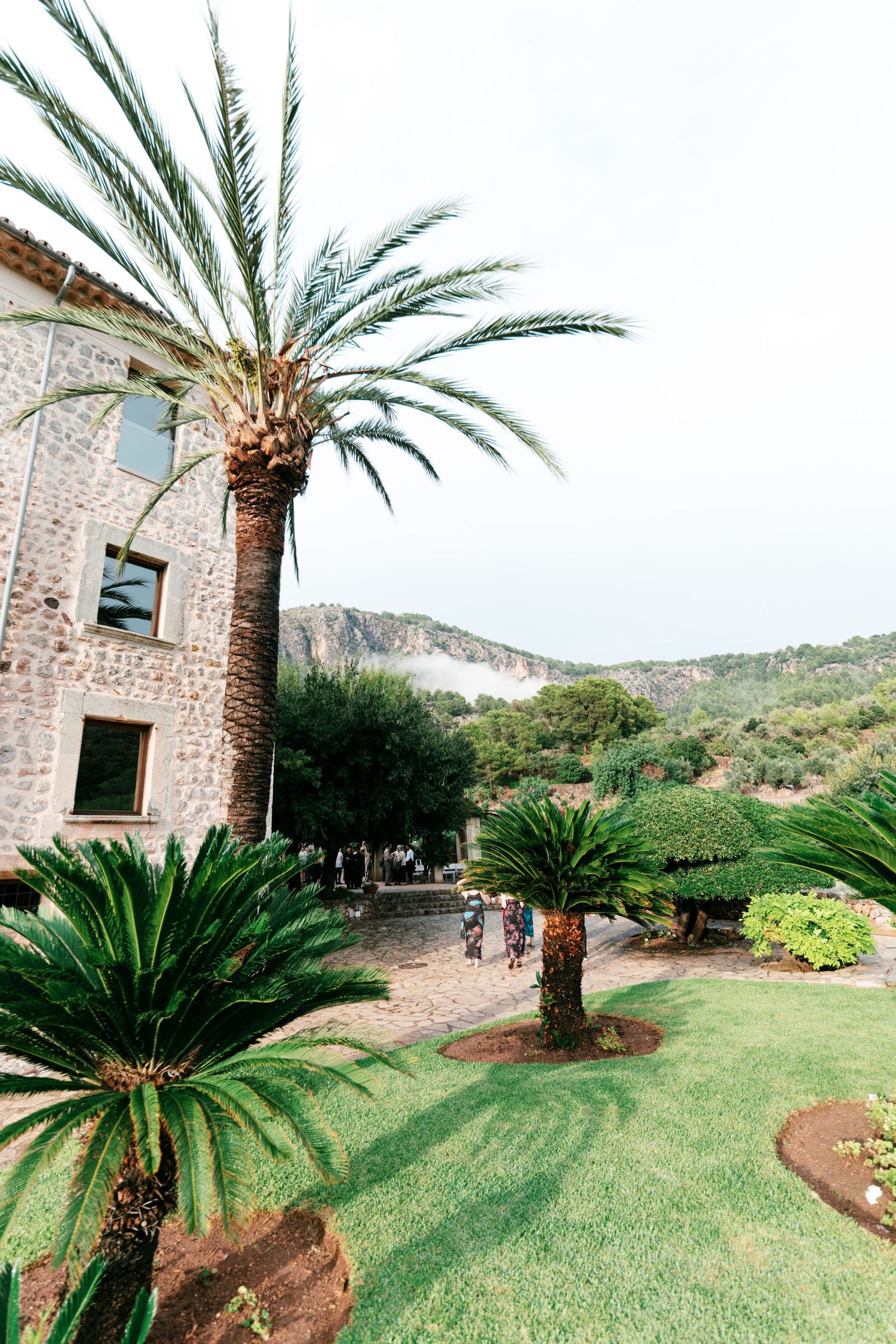 Stone villa with tall palm trees and a manicured garden beneath a rocky mountain backdrop on a sunny day.