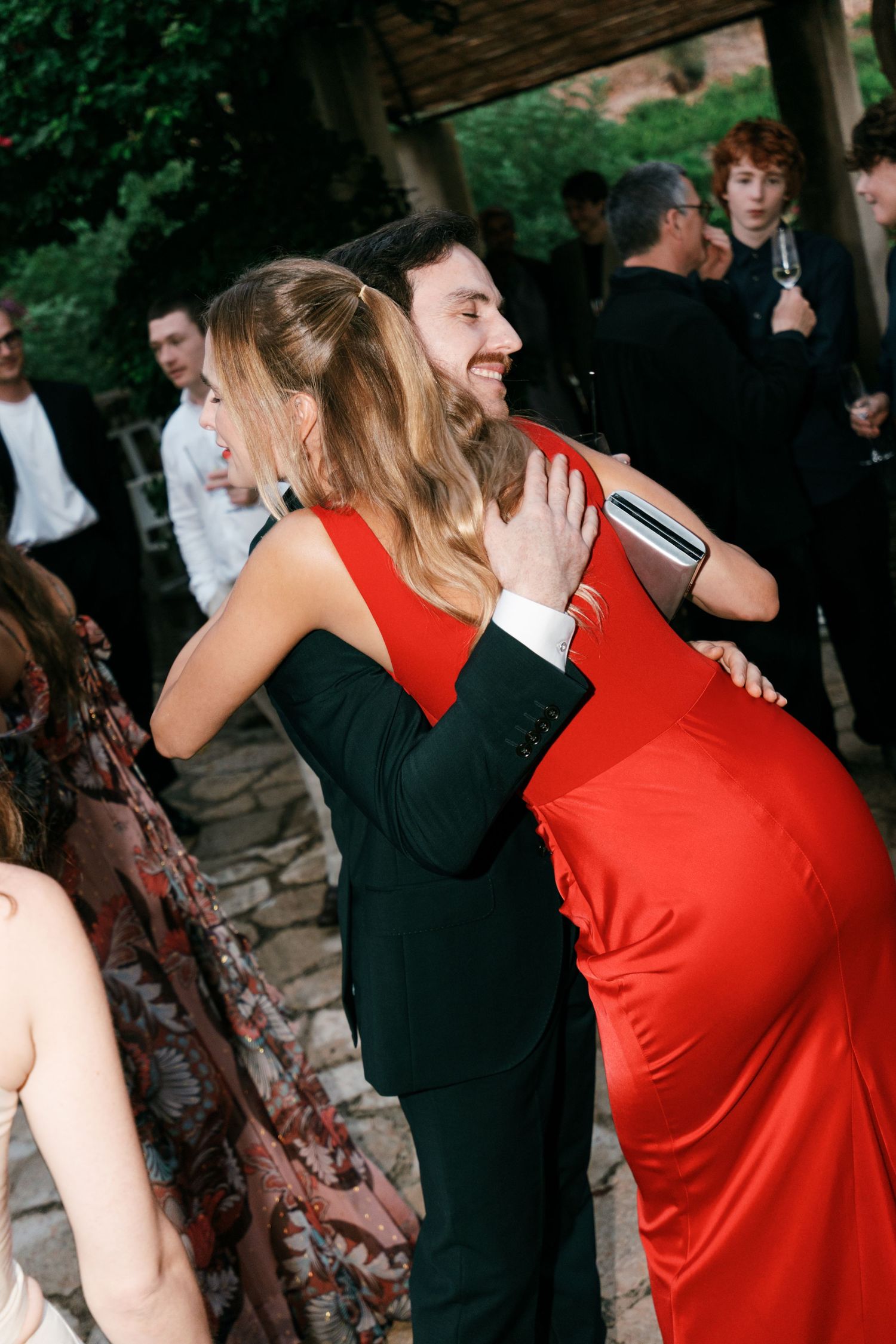 A woman in a red satin dress hugs a smiling man in a dark suit at an outdoor wedding reception while guests look on.
