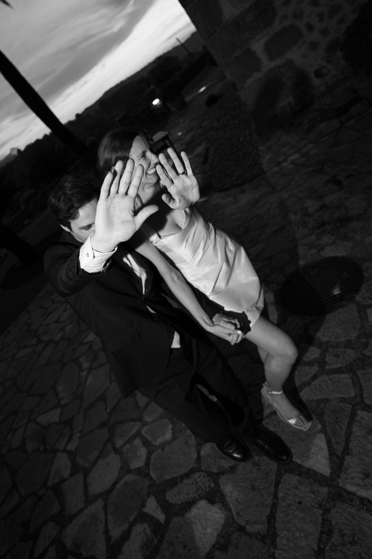 Black and white photo of a wedding couple playfully shielding their faces with raised hands on a cobblestone terrace.