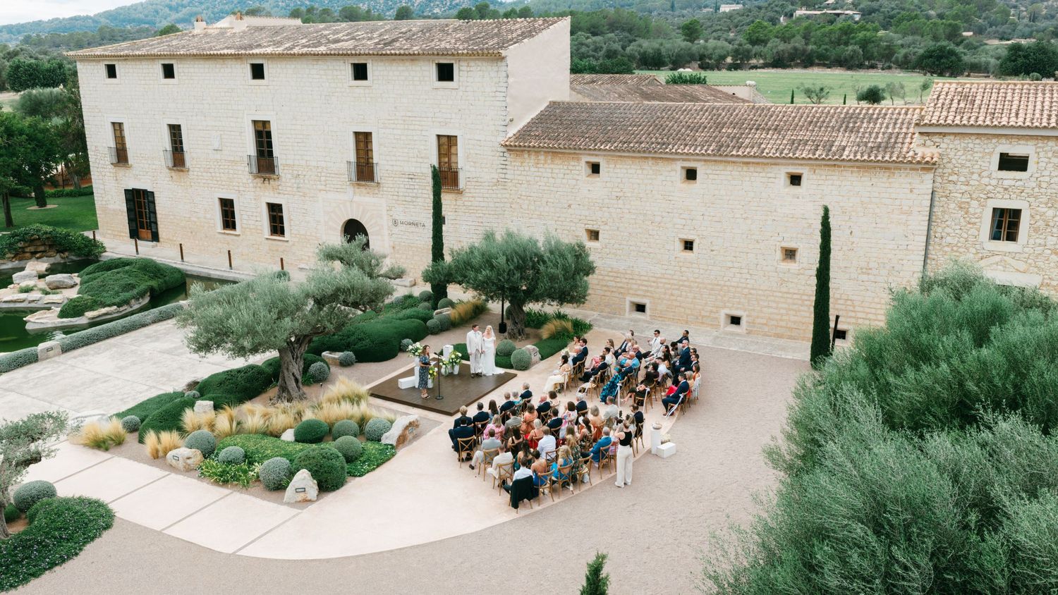 Aerial view of an outdoor wedding ceremony with guests seated in a landscaped garden courtyard beside a rustic stone Mediterranean estate.