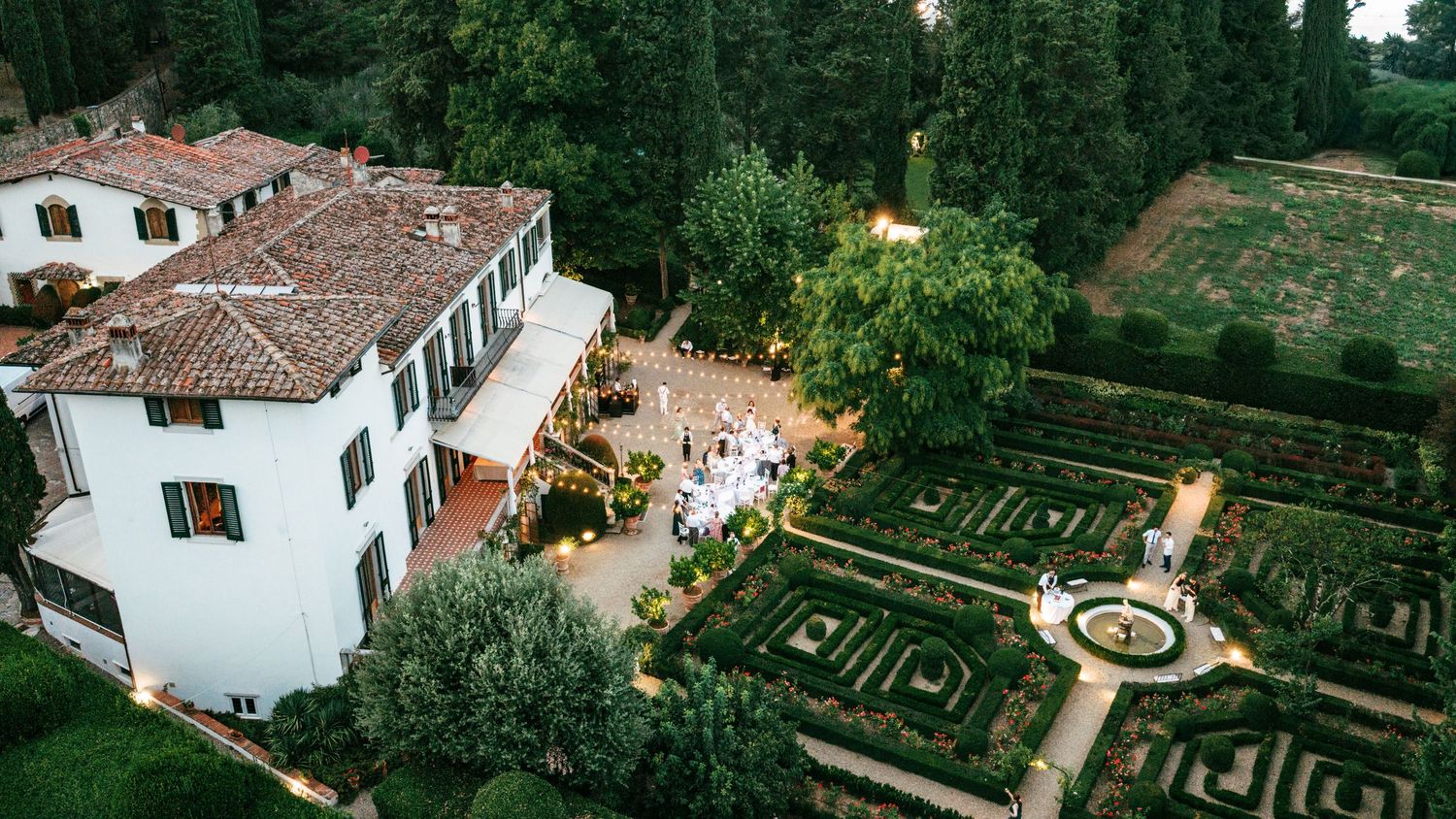 Aerial view of elegant Italian villa with manicured geometric gardens and outdoor wedding reception at dusk.