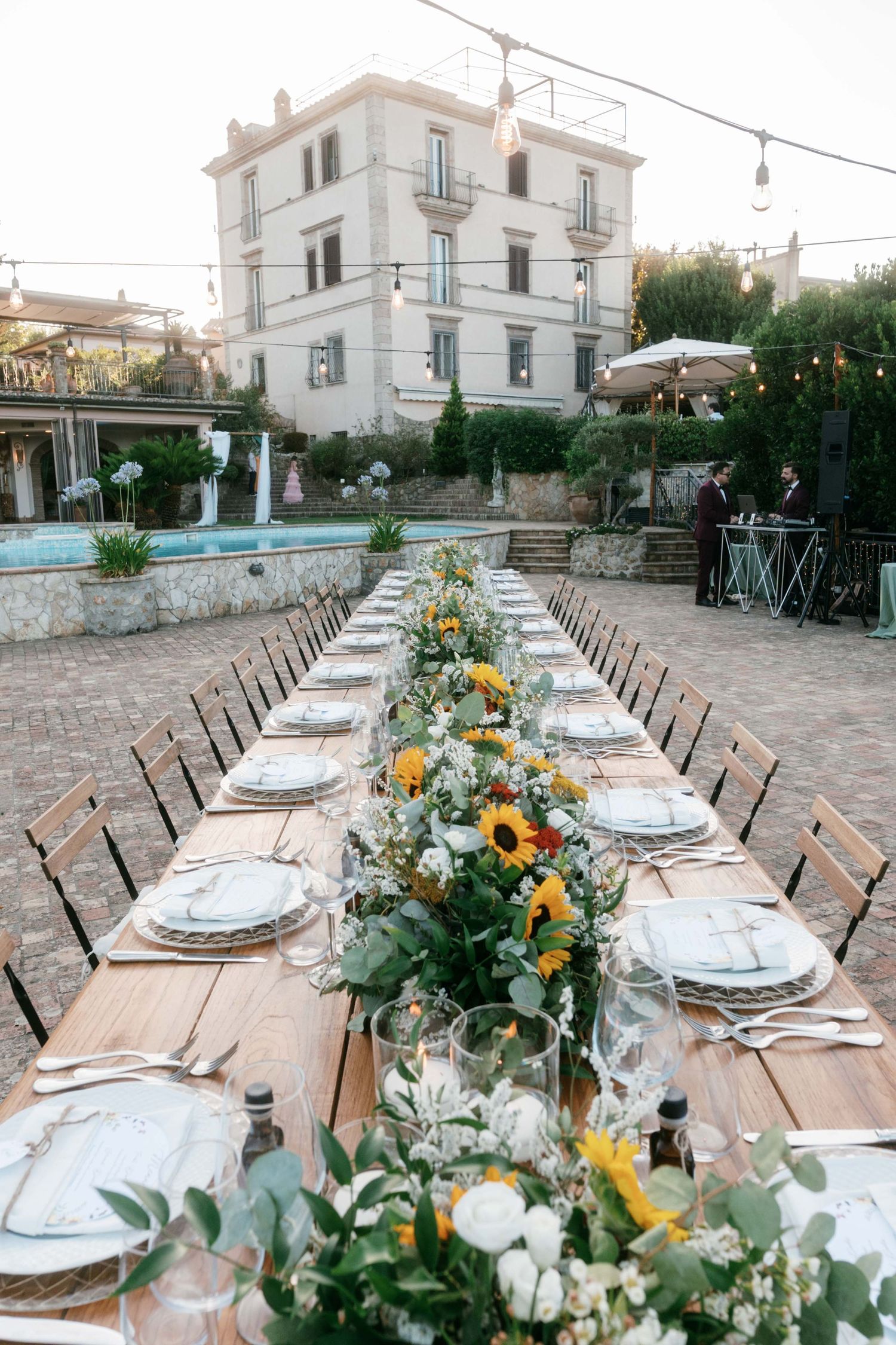 Long outdoor wedding reception table decorated with sunflowers and greenery in front of white Italian villa at dusk. Tuscany wedding photo