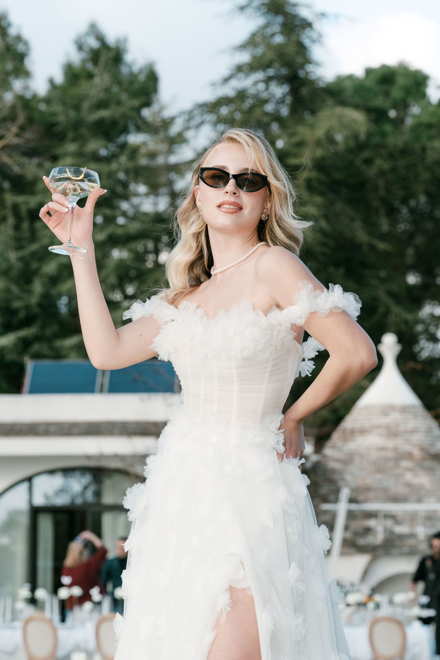 Stylish bride in white ruffled corset gown and sunglasses raising a champagne glass outdoors. Puglia wedding photographer, film