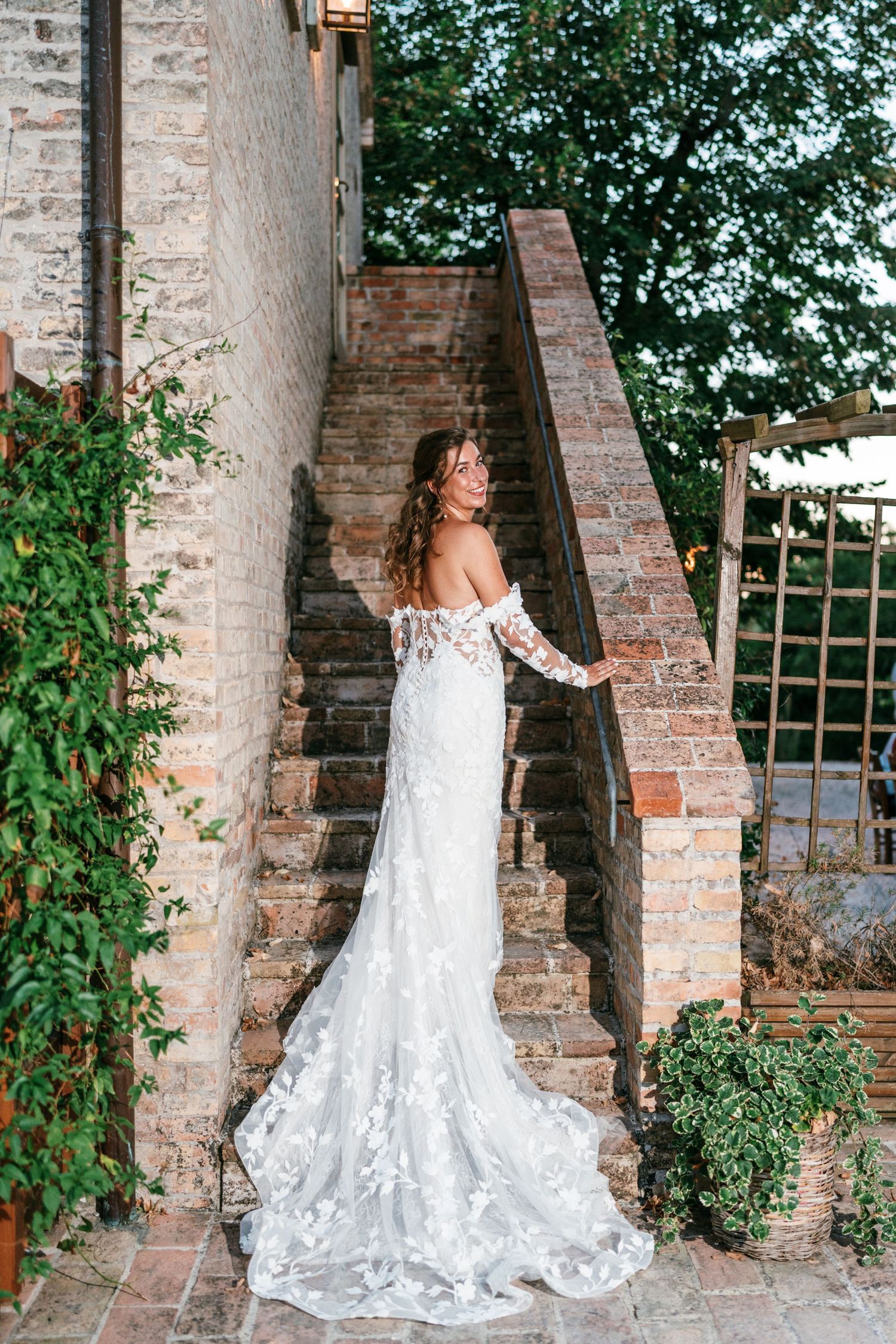 Bride in off-shoulder lace wedding gown with train poses on rustic brick staircase surrounded by greenery.