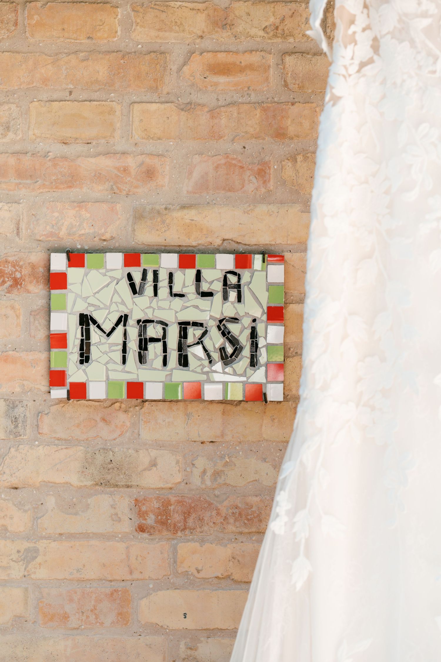 Mosaic tile sign reading Villa Marsi mounted on a brick wall beside a white wedding dress.