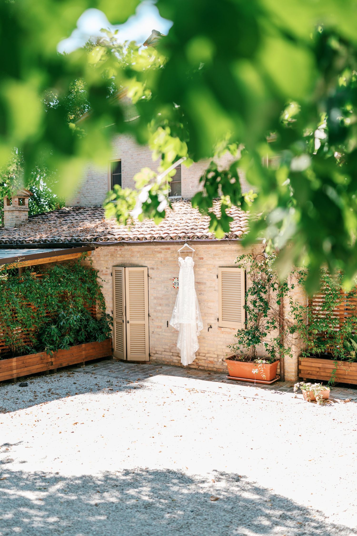 White wedding dress hanging outside a rustic brick Italian villa with wooden shutters, terracotta roof tiles, and potted plants in sunlight.