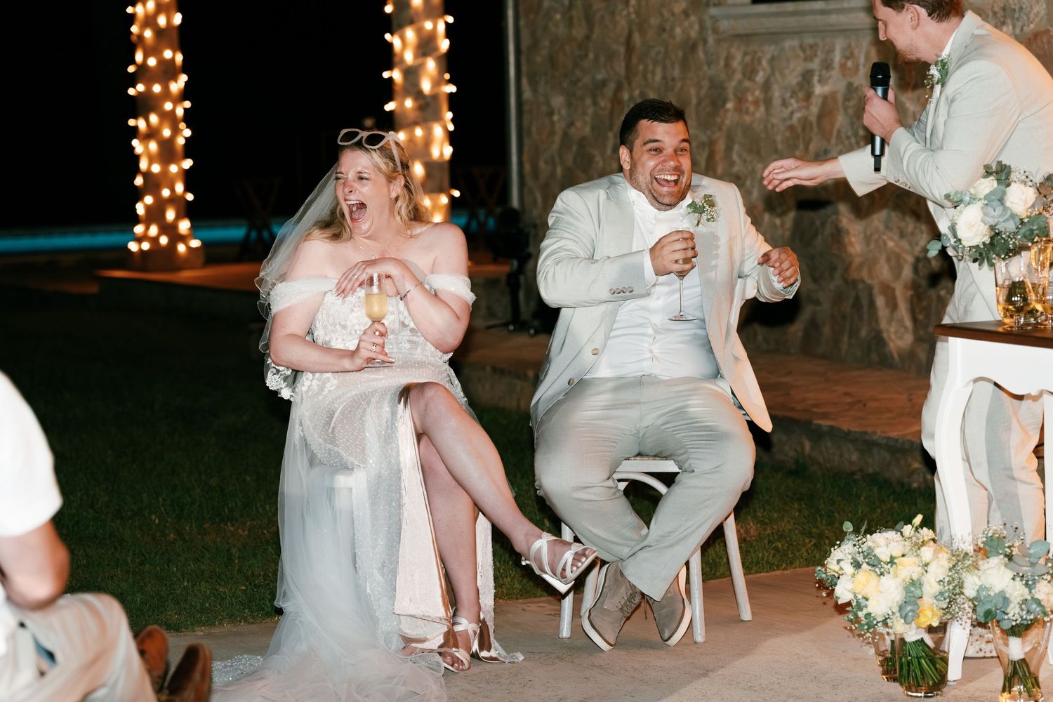 Bride and groom laugh joyfully while seated during a fun wedding reception game at night. Mallorca wedding photographer, filmmaker