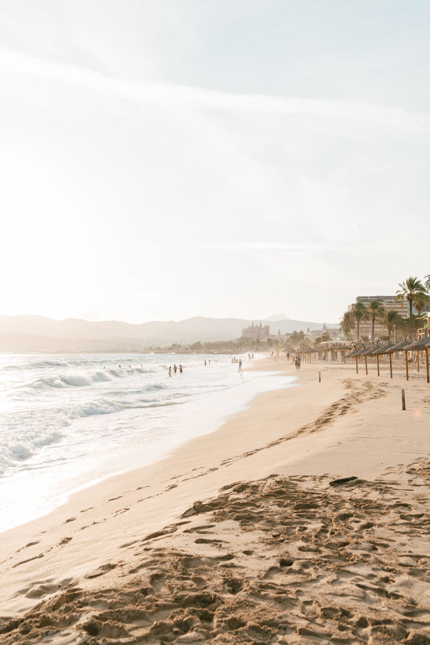 Sun-kissed sandy beach with gentle waves, thatched umbrellas, and mountains in the background during golden hour. Mallorca wedding filmmaker