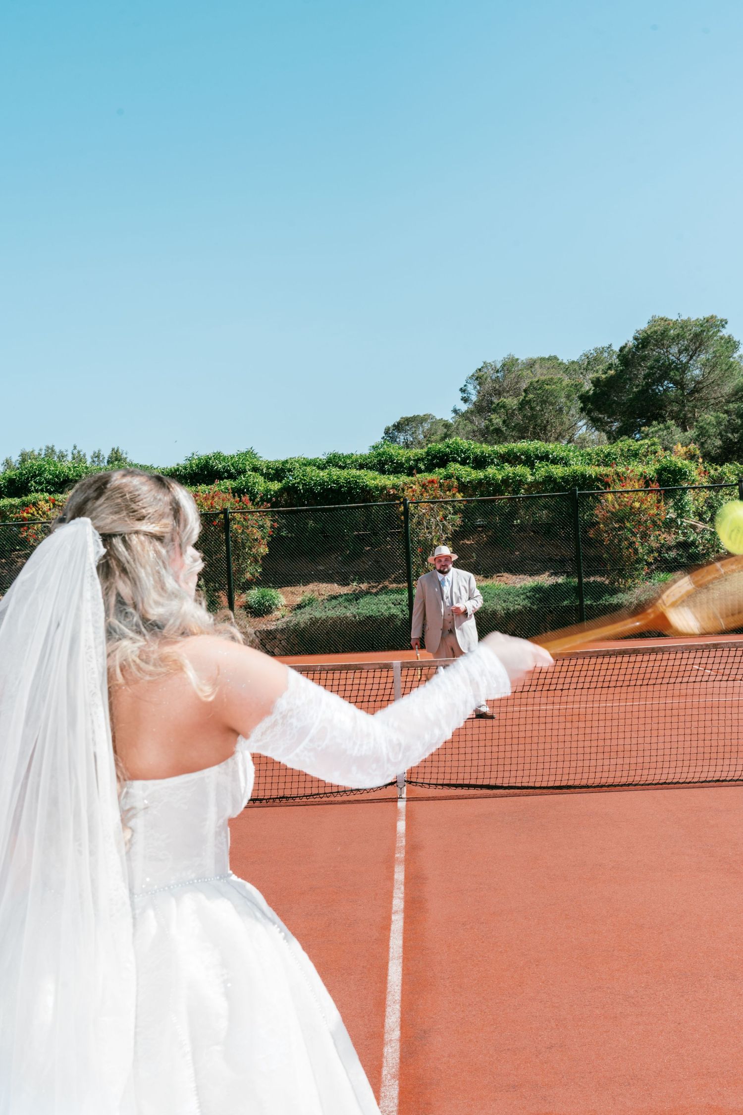 Bride in white gown and veil plays tennis on clay court while groom stands at the net on sunny day.