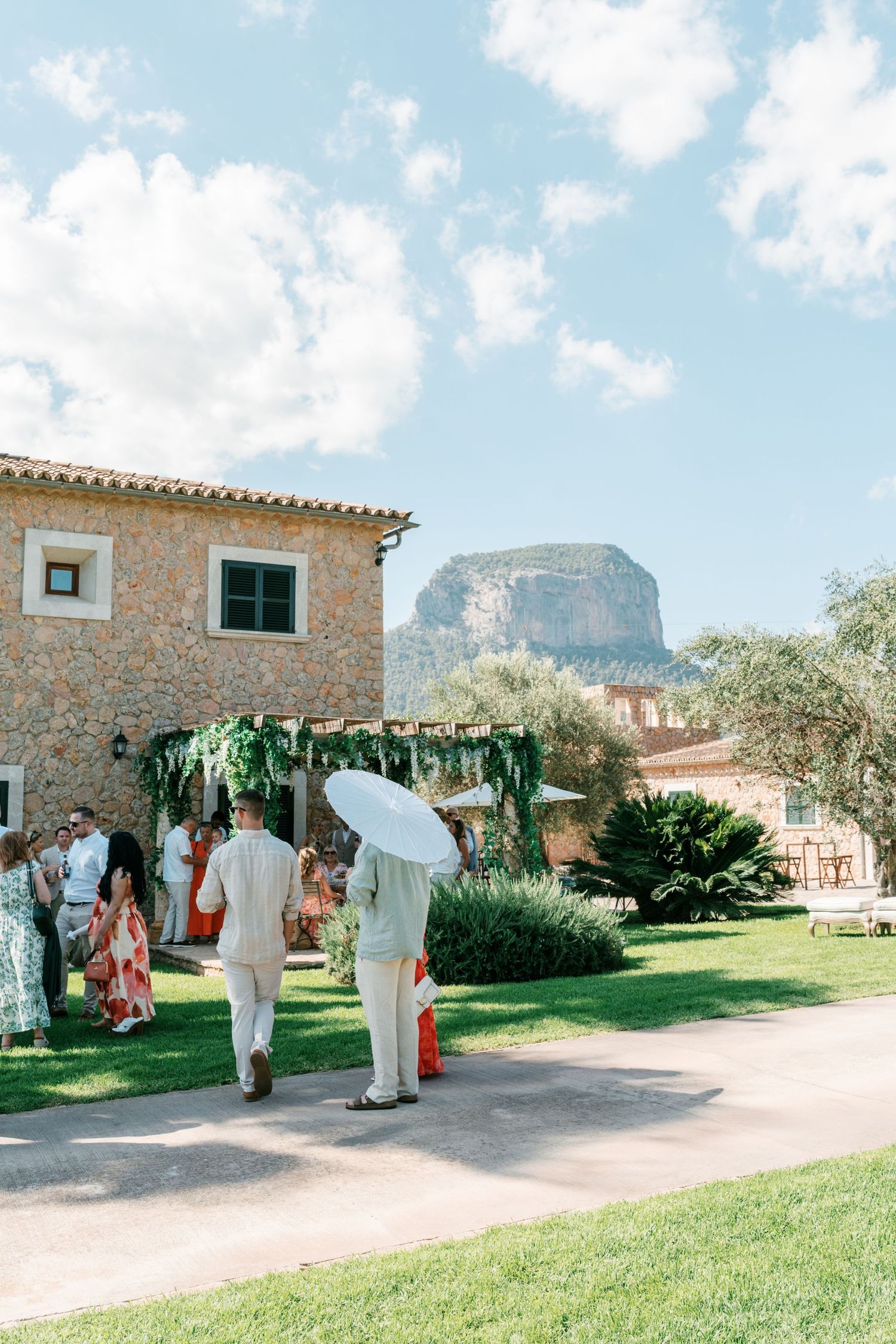Two guests walk toward a rustic stone villa wedding venue with a dramatic rocky mountain backdrop under blue skies.