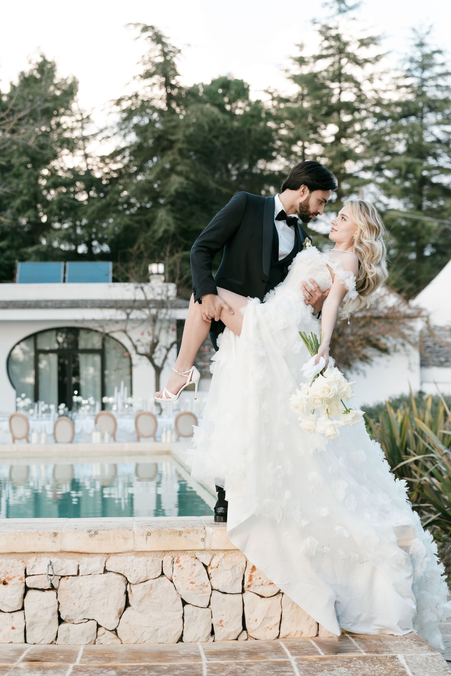 Groom dipping bride in layered white gown beside a pool at an elegant outdoor wedding venue. Mallorca wedding photographer, filmmaker