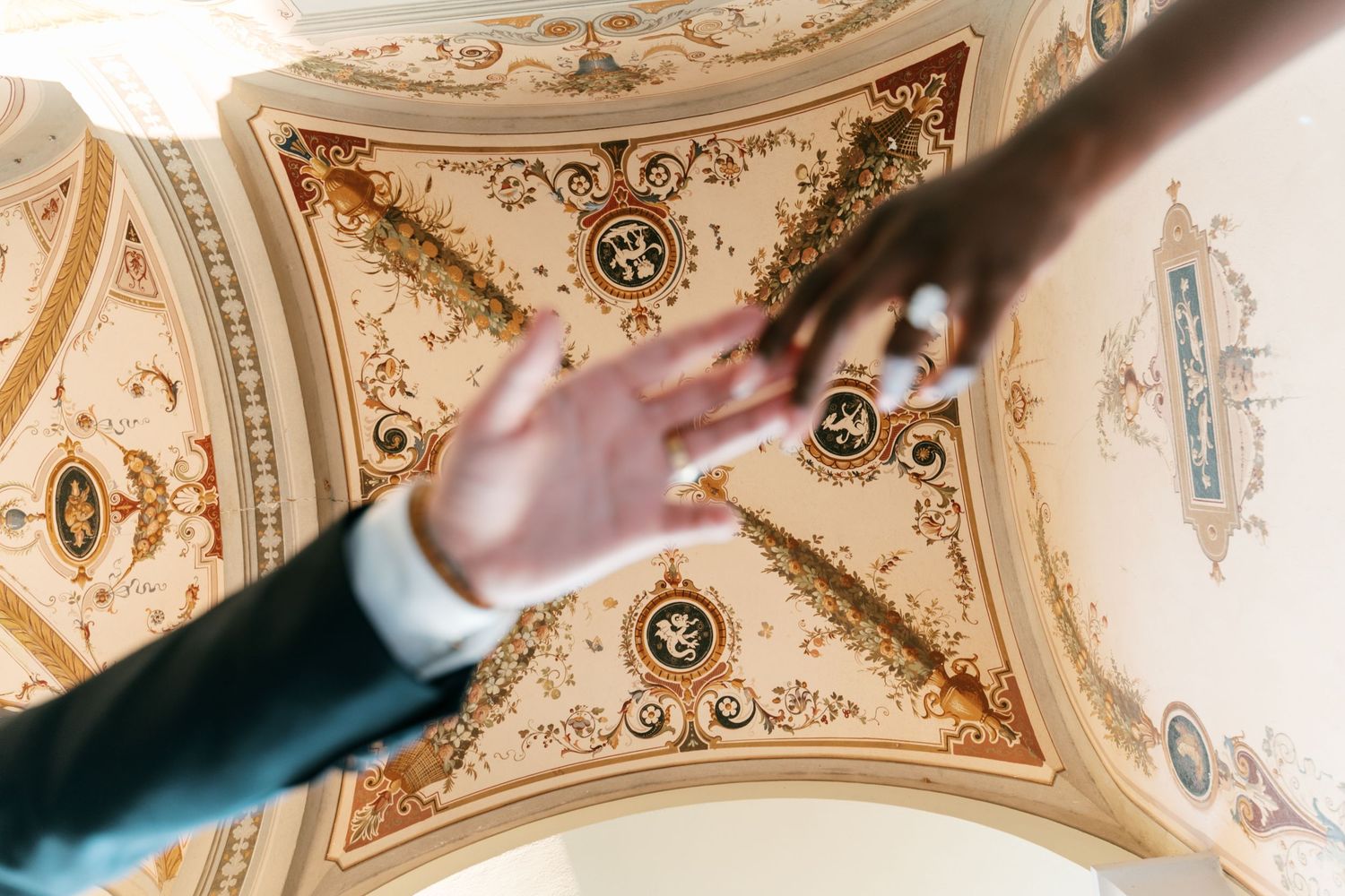 Two hands reaching toward each other beneath an ornate frescoed ceiling with gold decorative medallions, as seen from below at a wedding.