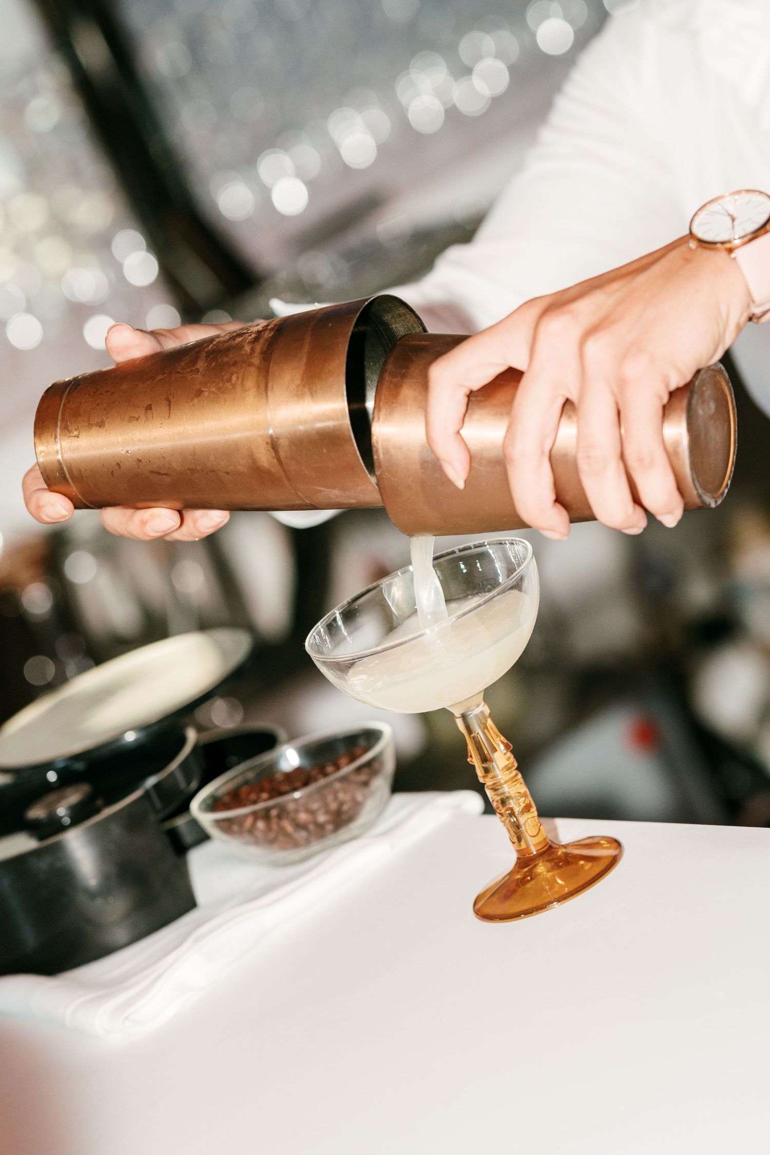 Bartender pouring a creamy cocktail from a copper shaker into a vintage amber coupe glass, with a bowl of coffee beans nearby.