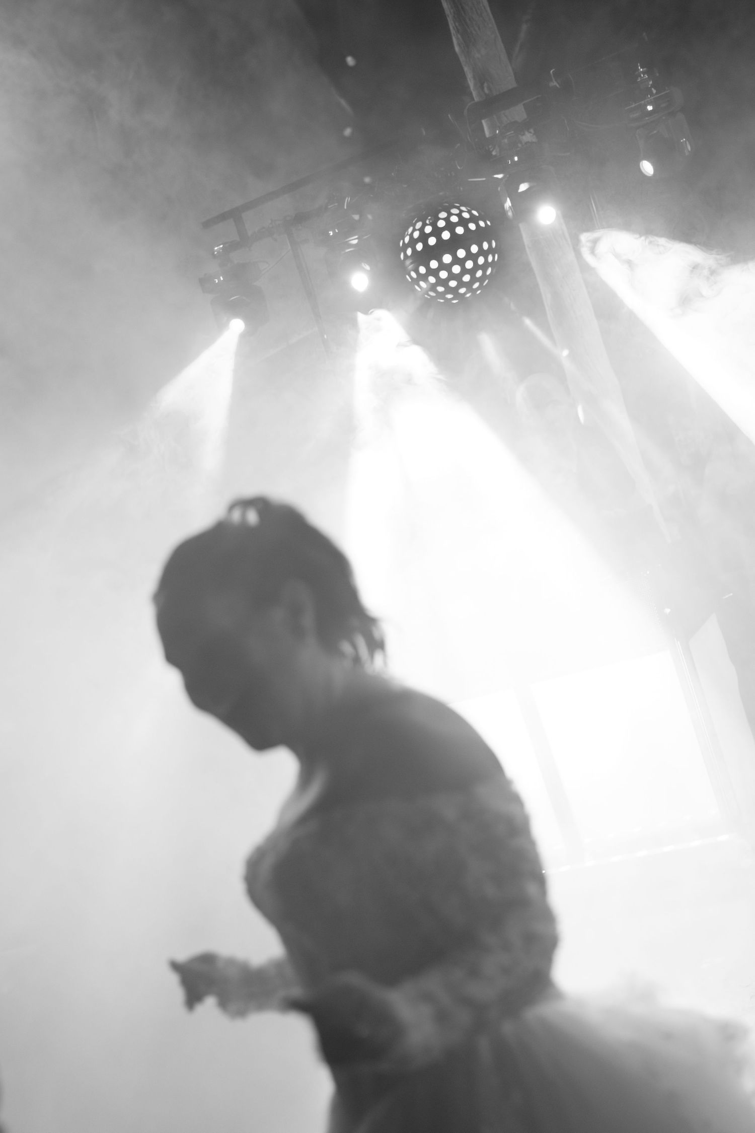 Black and white photo of a bride in a lace gown dancing beneath a disco ball and hazy stage lights at a wedding reception.