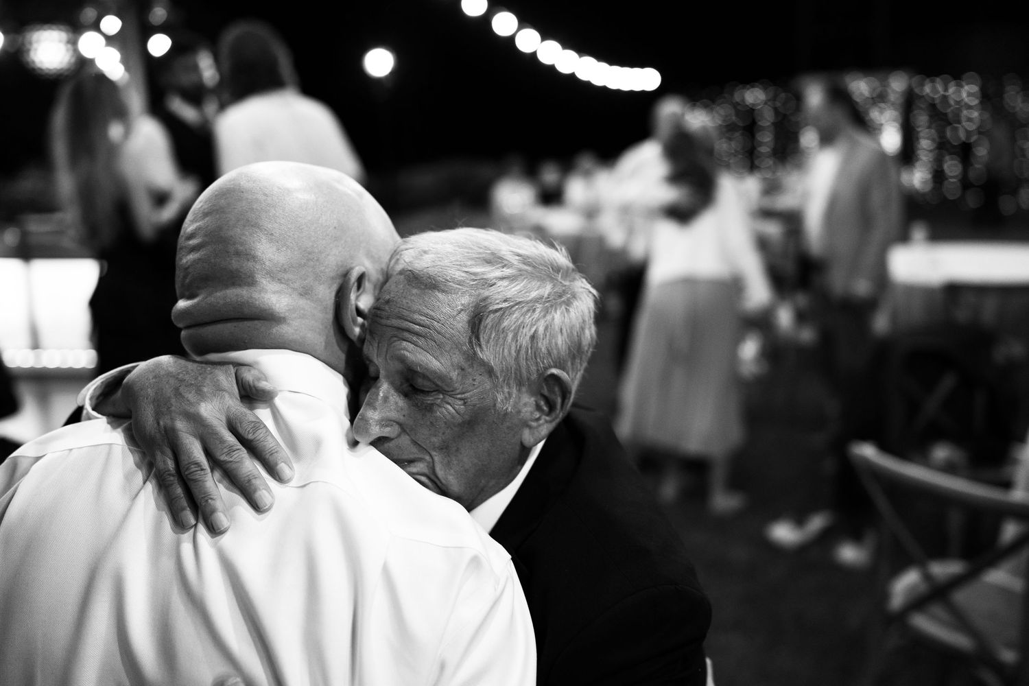 Two men share a heartfelt embrace at an evening celebration, with guests and string lights softly blurred behind them in black and white.