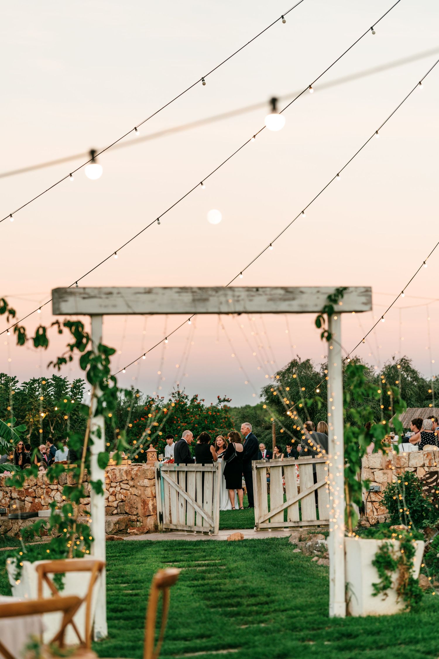 Wedding guests gather at dusk beneath string lights near a rustic wooden gate and a vine-covered pergola in a garden venue.