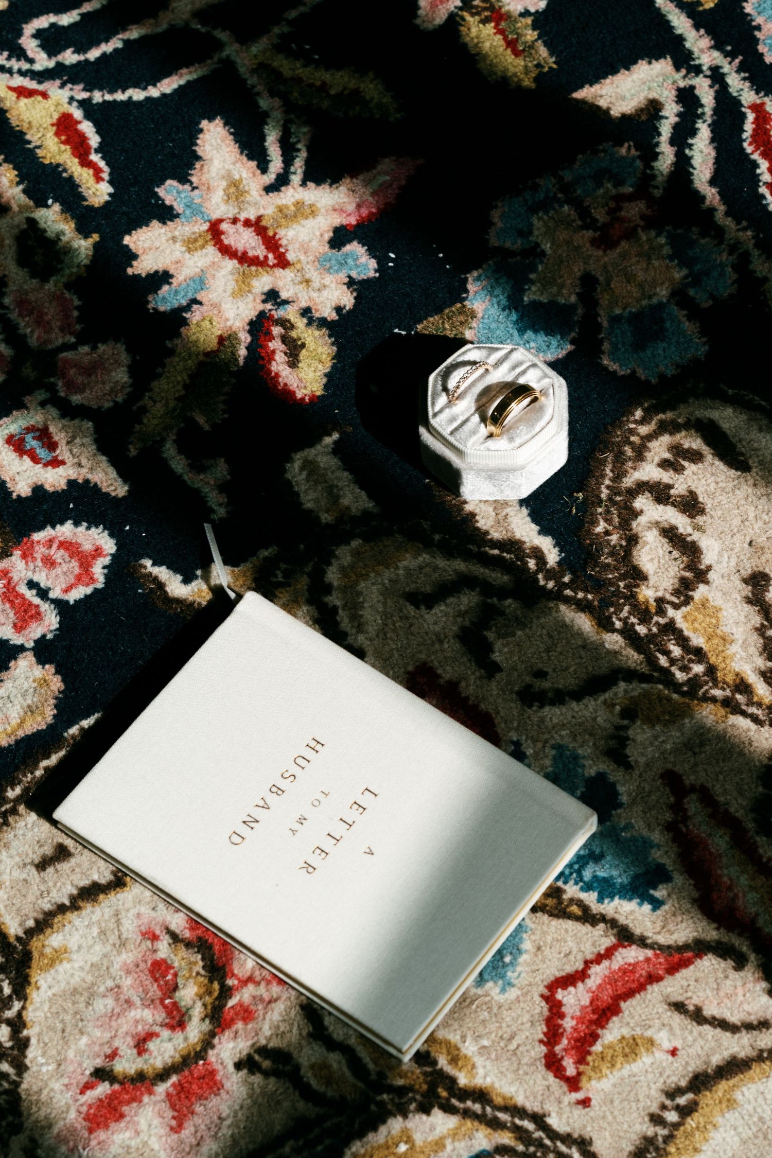Wedding rings in a white hexagonal ring box beside a card reading A Letter to My Husband on an ornate floral Persian rug.