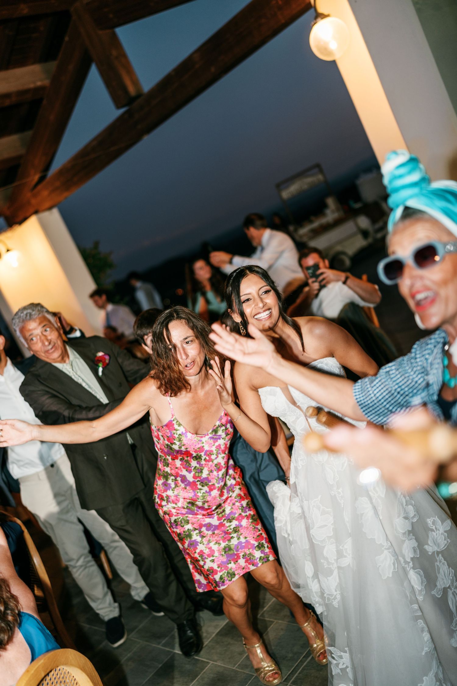 Guests dance joyfully at an outdoor evening wedding reception, with a bride in a white lace gown laughing beside a woman in a floral dress.